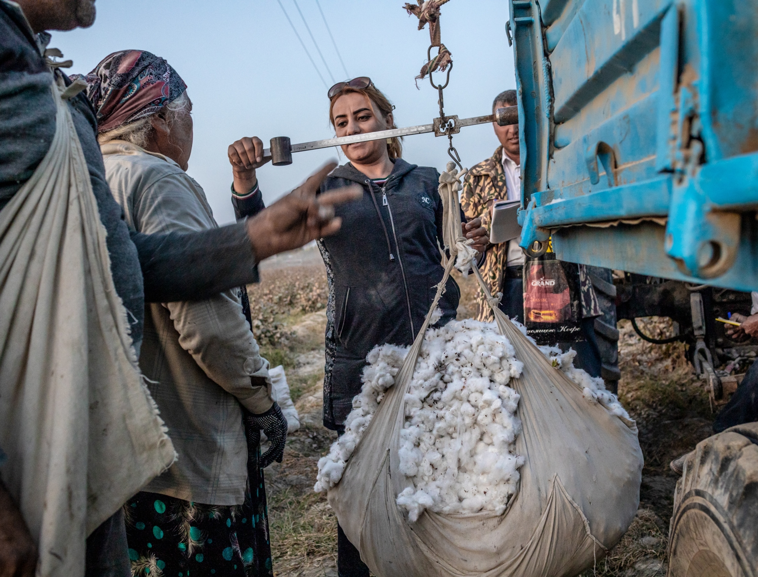 Woman by the torque colored dump truck using balance scales to weight a bag of cotton. Three more people watching her doing it.