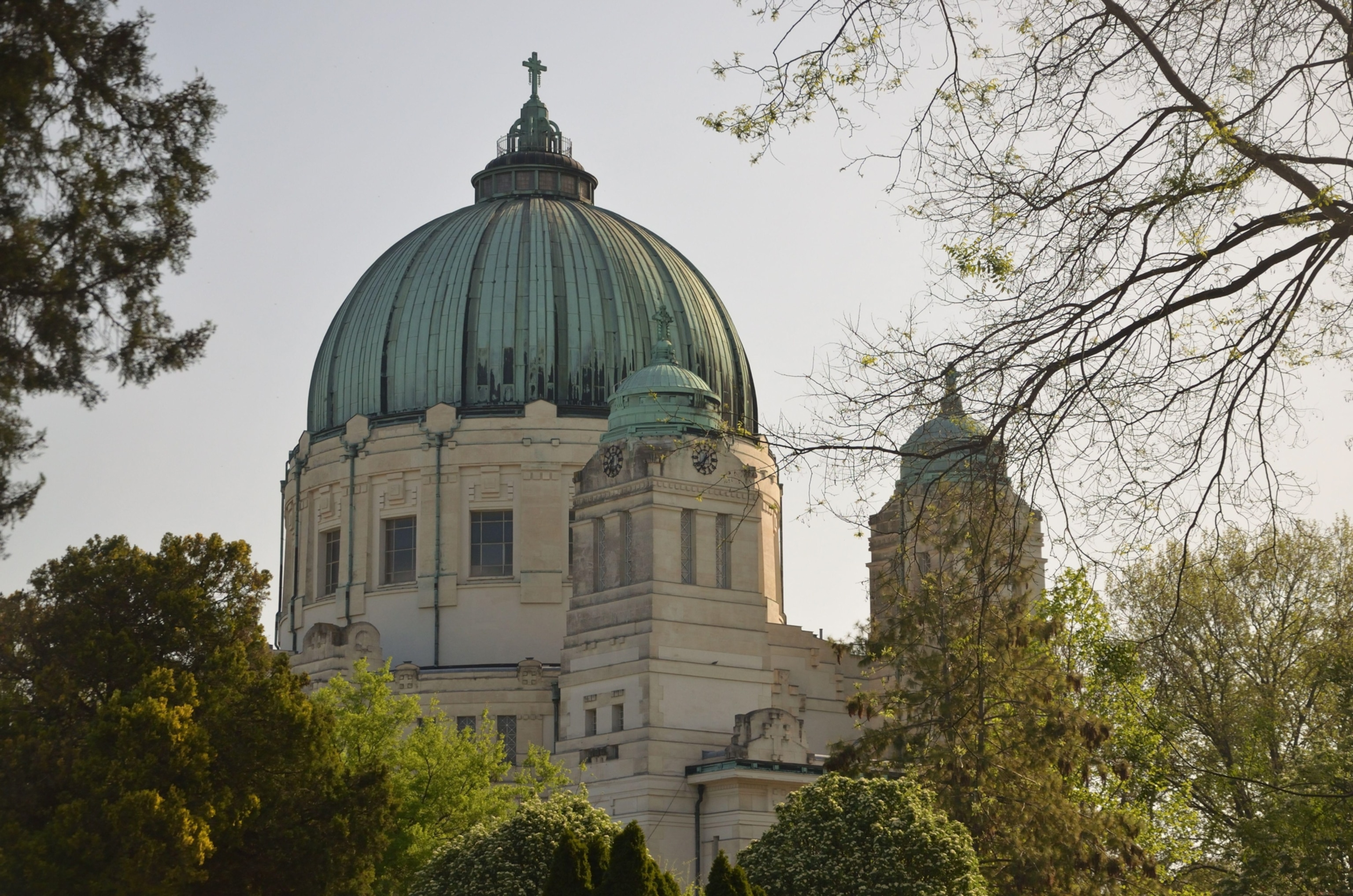 A large domed church surrounded by trees stands above a historic cemetery, with a cross visible at the top of the copper-colored dome.