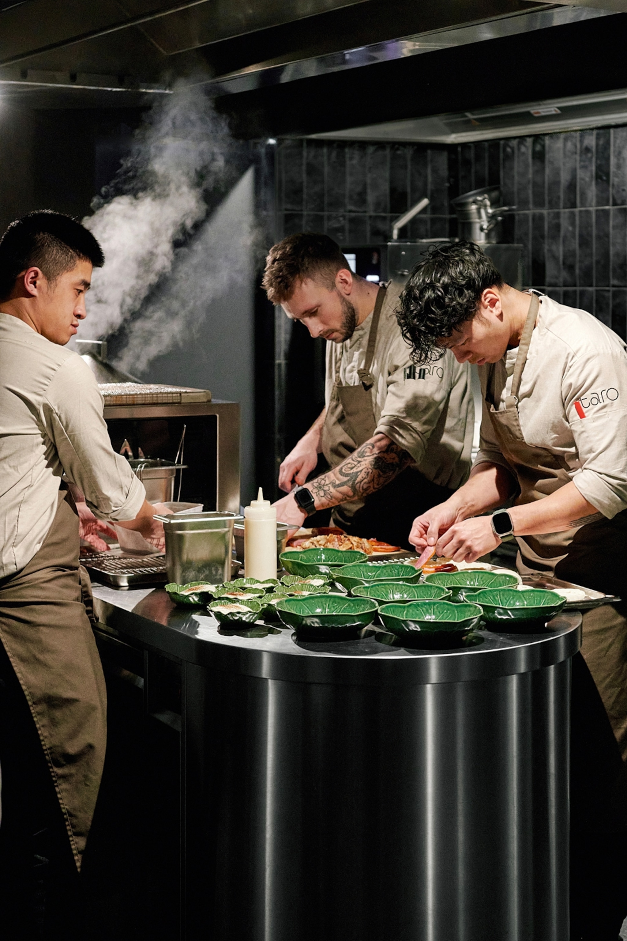 Three chefs perched around a counter in a kitchen, finishing up plates.