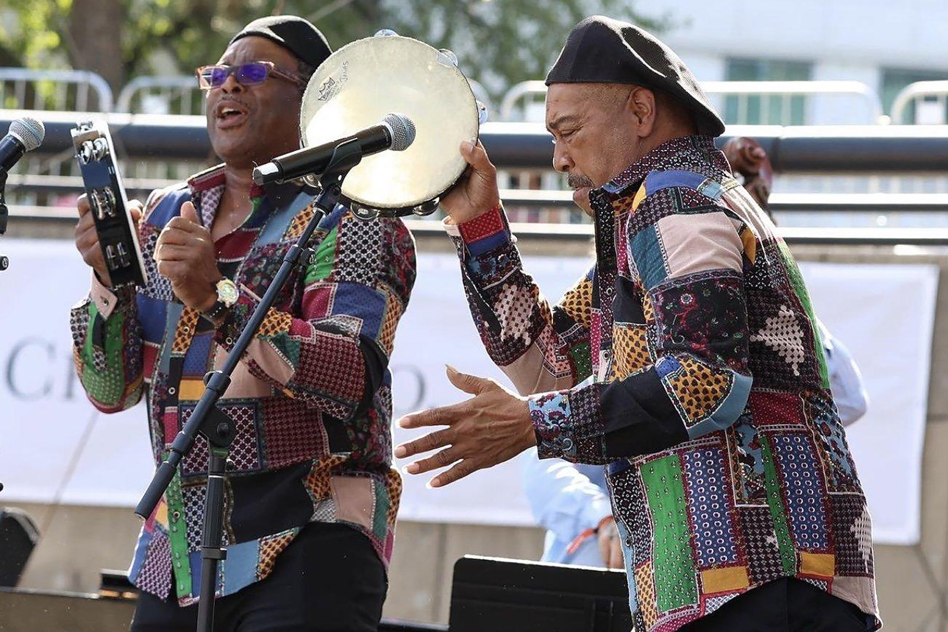 Two musicians in quilted patterned shirts playing the tambourine and dancing with microphones stood in front of them