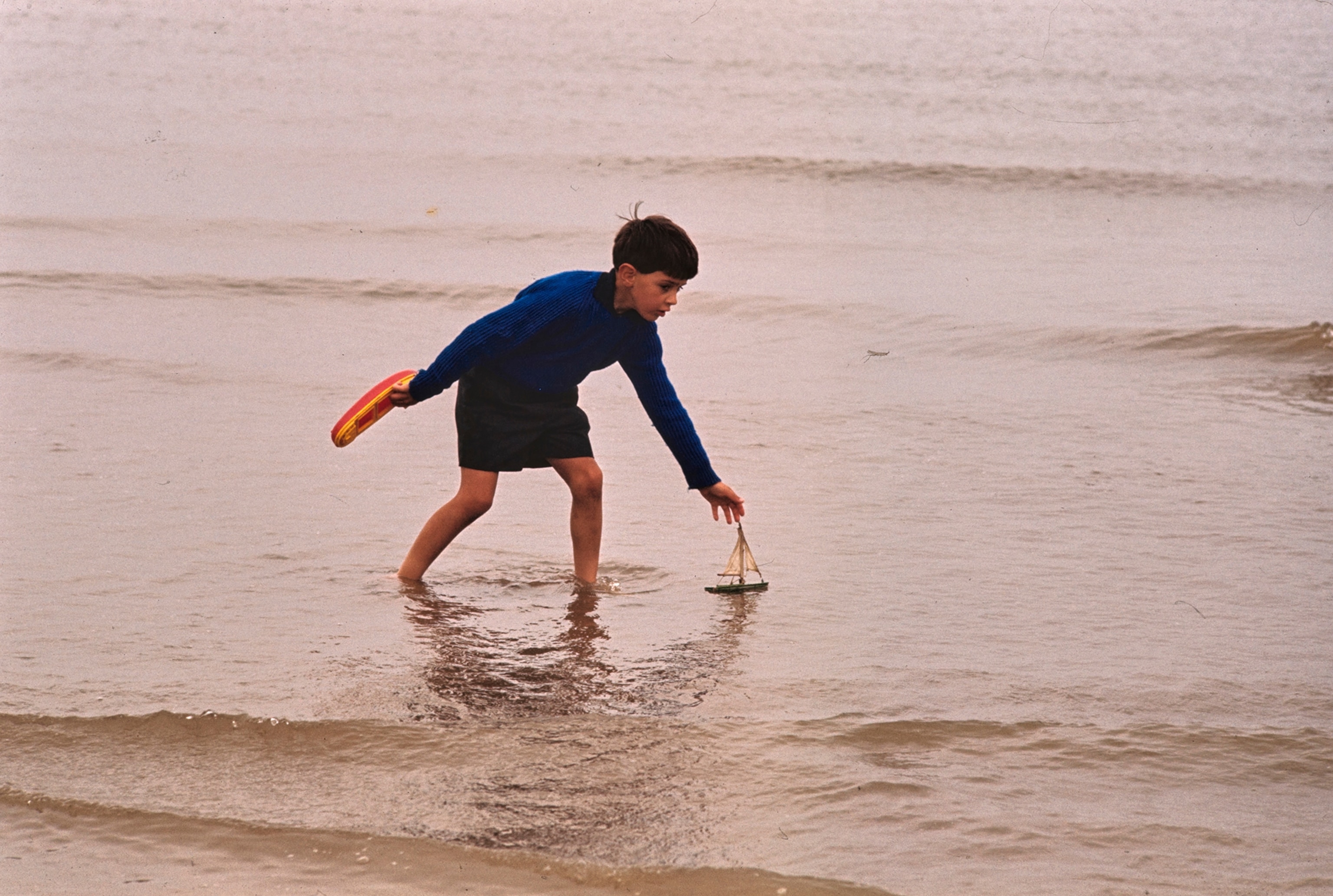 Boy plays with model boats in surf.