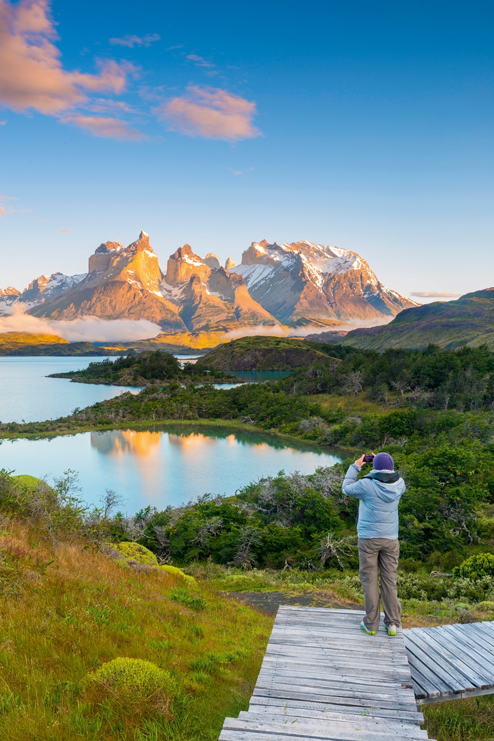 A landscape shot of a hiker taking a picture or a lake-dotted flat plaine with snow-capped peaks in the background.