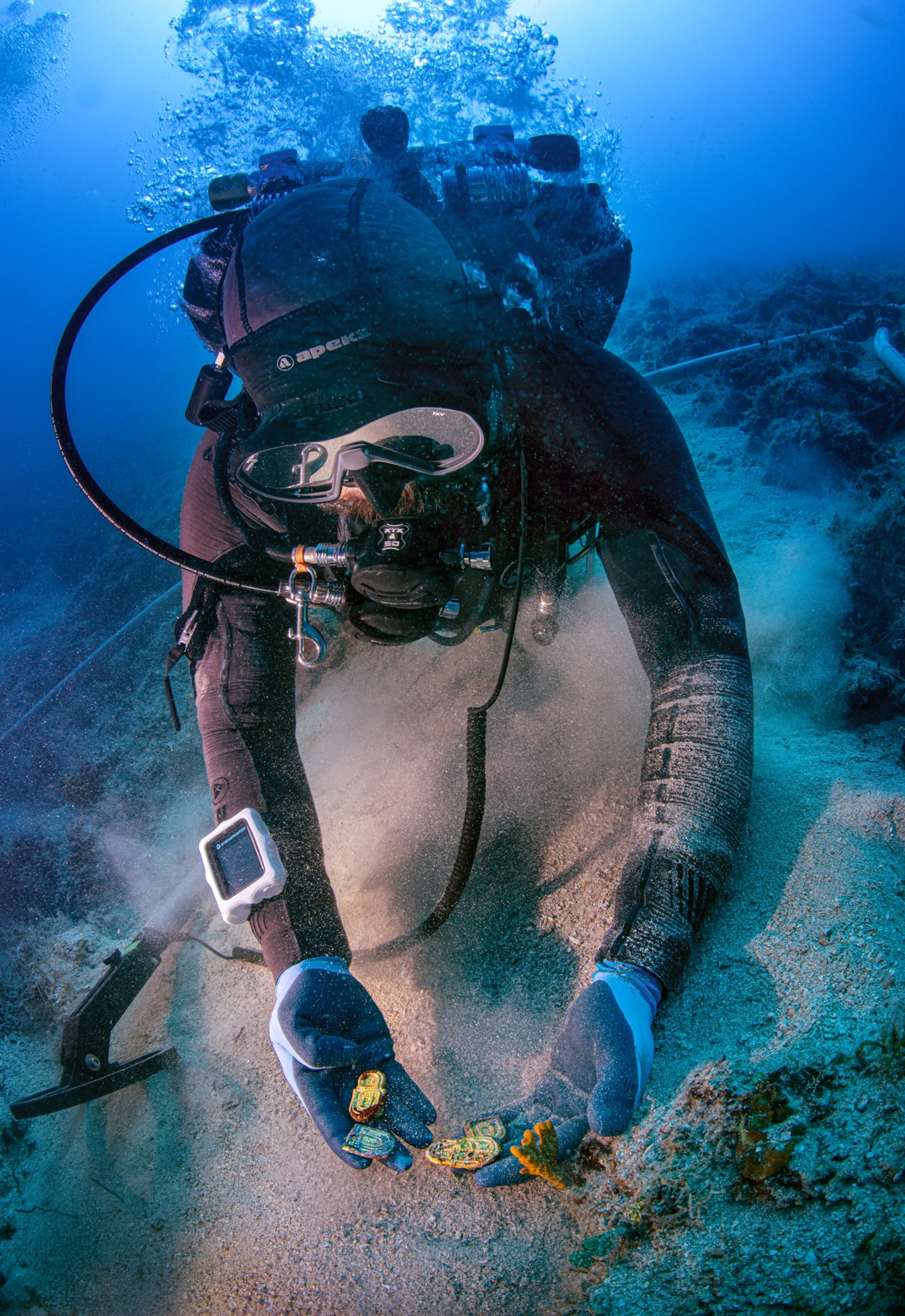 A diver underwater on the sea floor holds out various items that have been found.