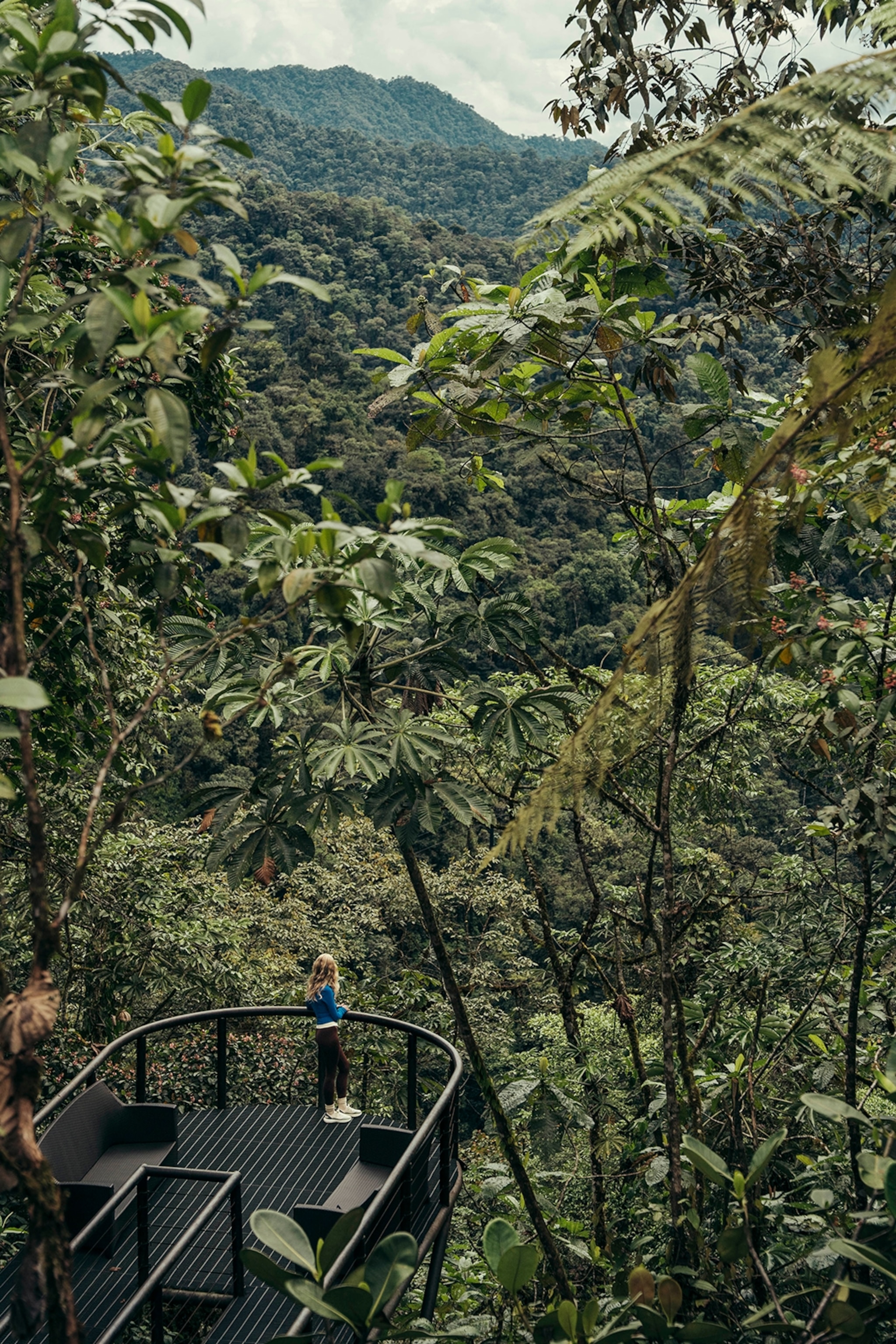 A young woman standing on the edge of a steel balcony in a tropical forest with dense vegetation and hills stretching out in front.