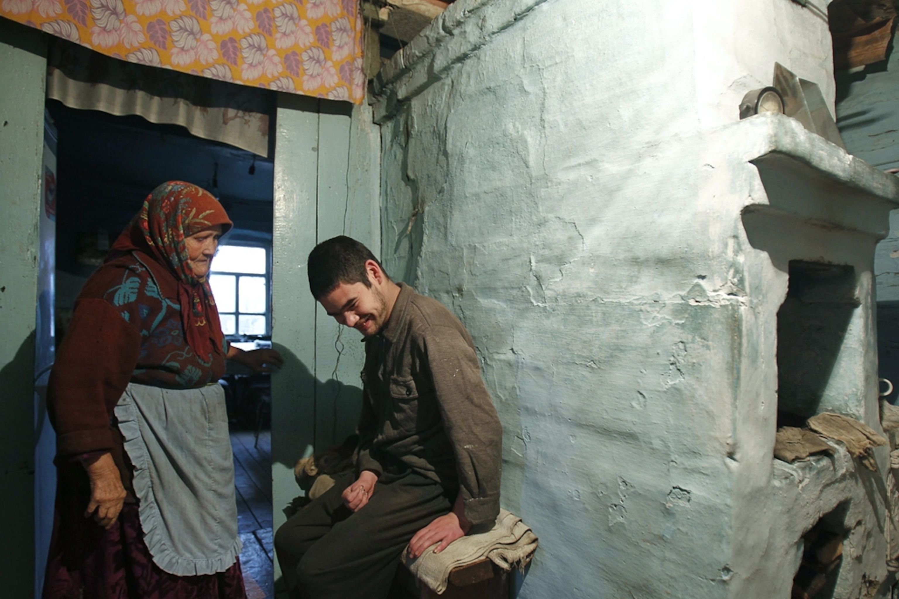 A young man suffering from cerebral palsy sits near the hearth of his family home while an elderly woman looks on.