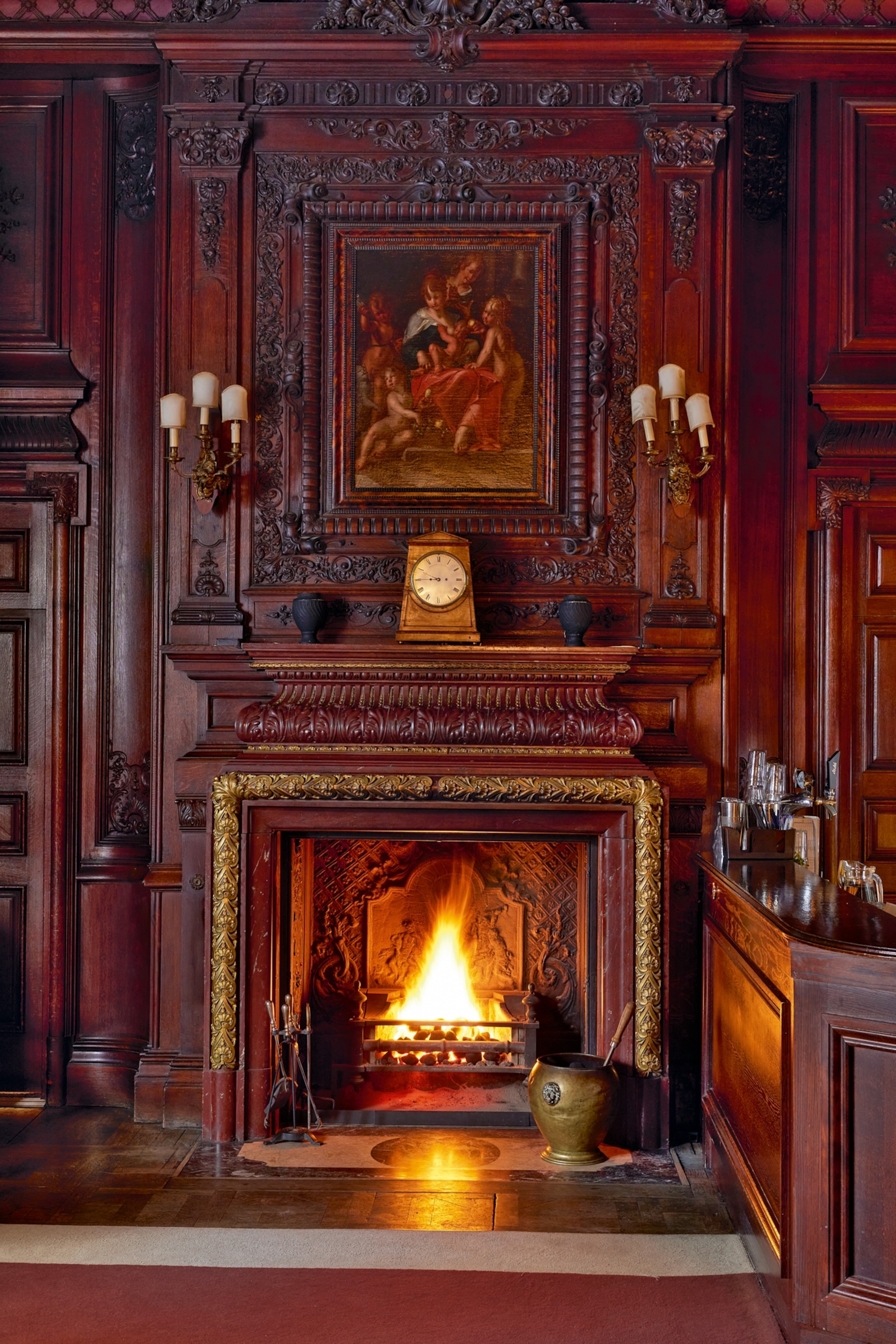 Ornate wooden fireplace with intricate carvings and a golden-framed fire. Above, a classic painting and a clock, flanked by elegant wall sconces, create a warm, luxurious atmosphere.