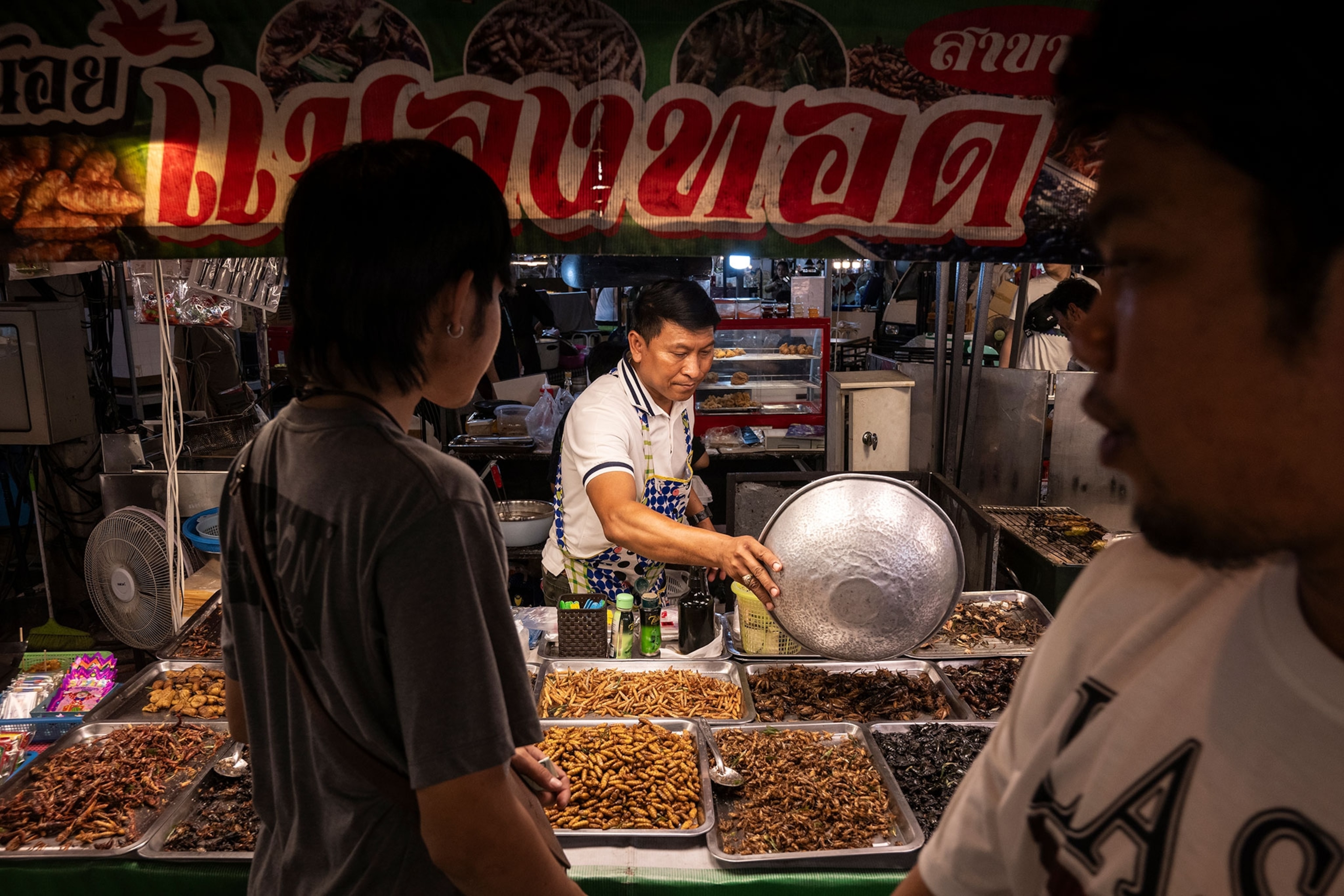 A stand at a night market is filled with a variety of trays of edible insects. A man working behind the stand is filling a tray while two people crossed in front.