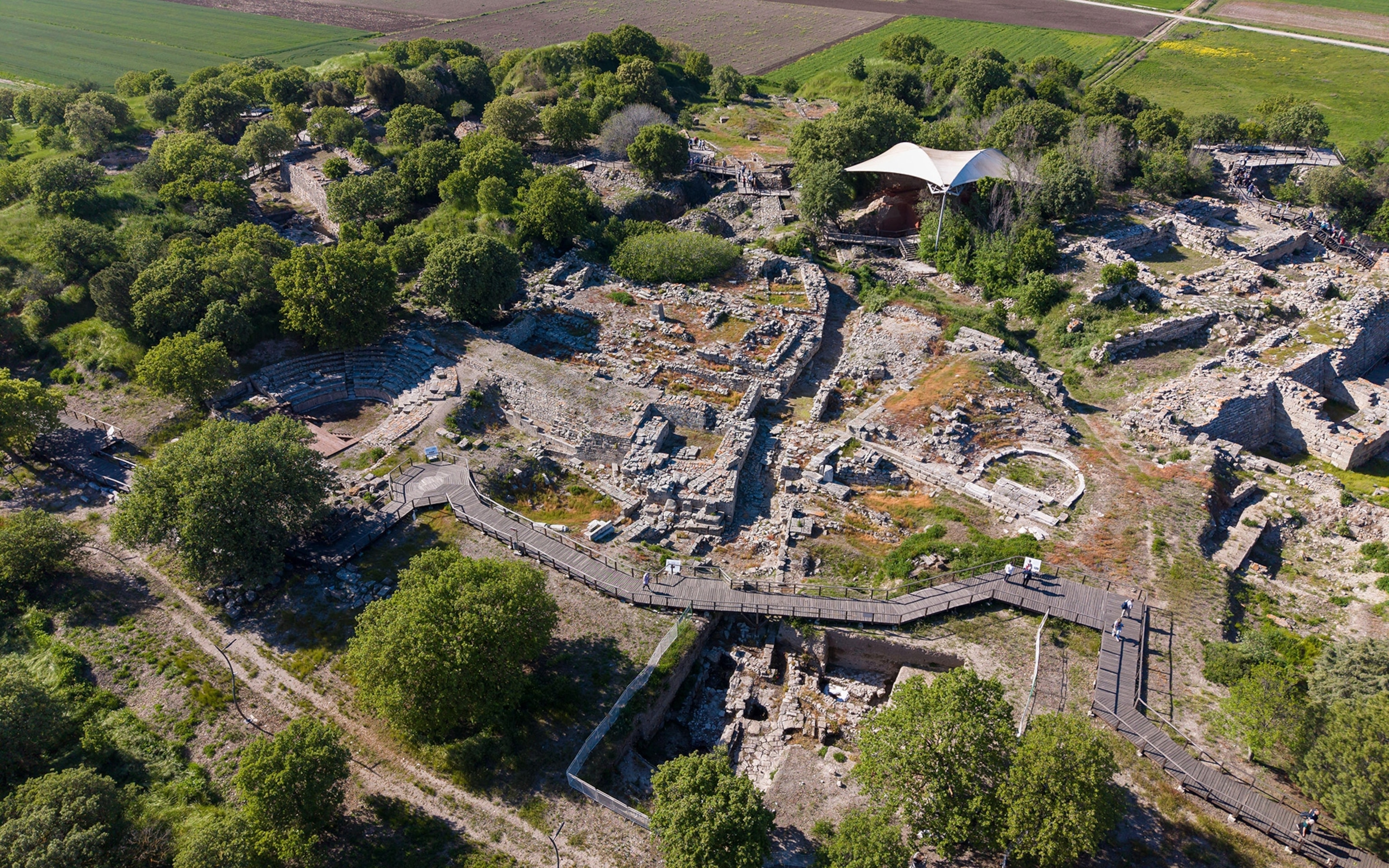 Aerial view of an archaeological site surrounded by green trees and fields, a wooden walkway along front