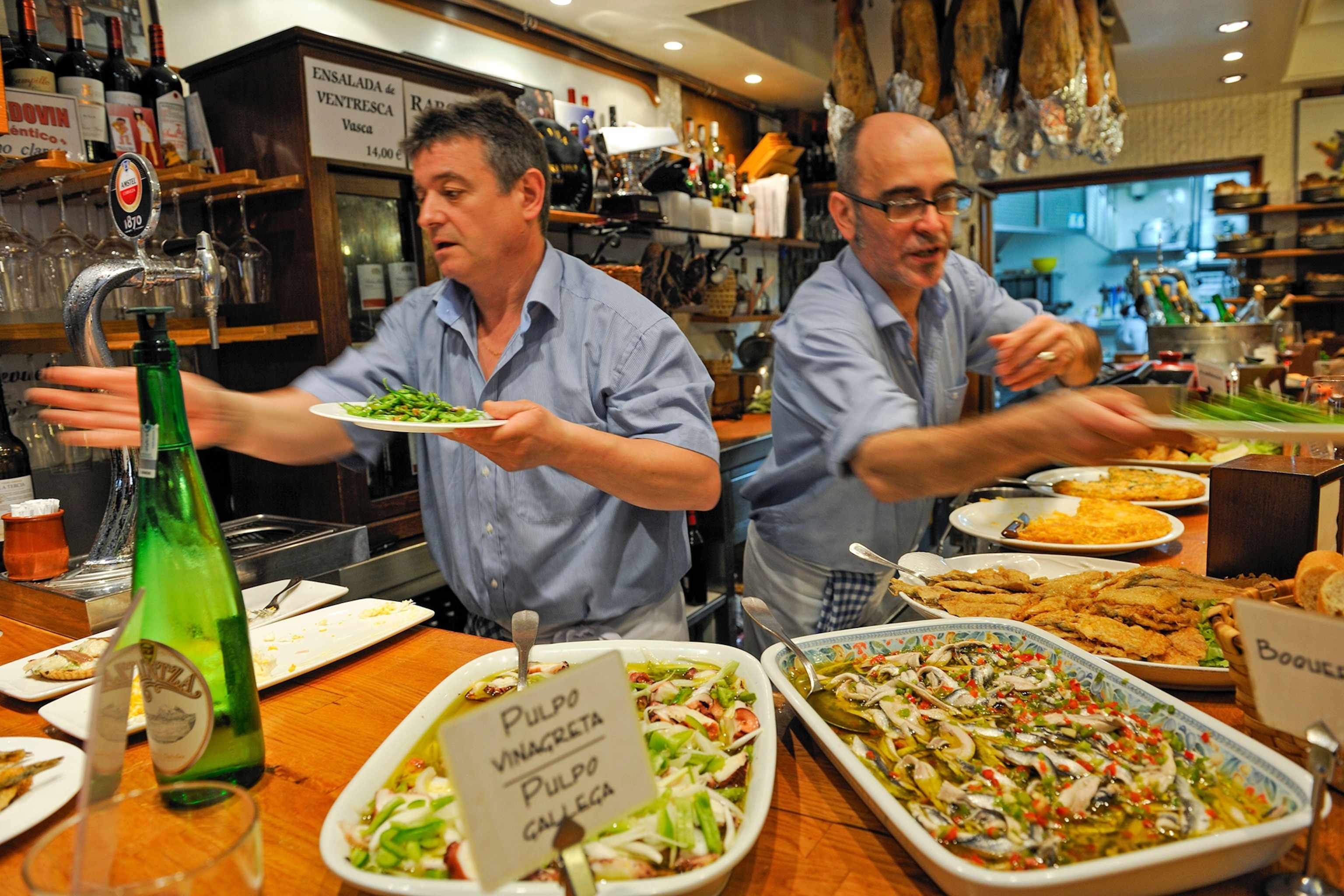 Two men behind a bar decked with large platters of food dynamically serving guests.