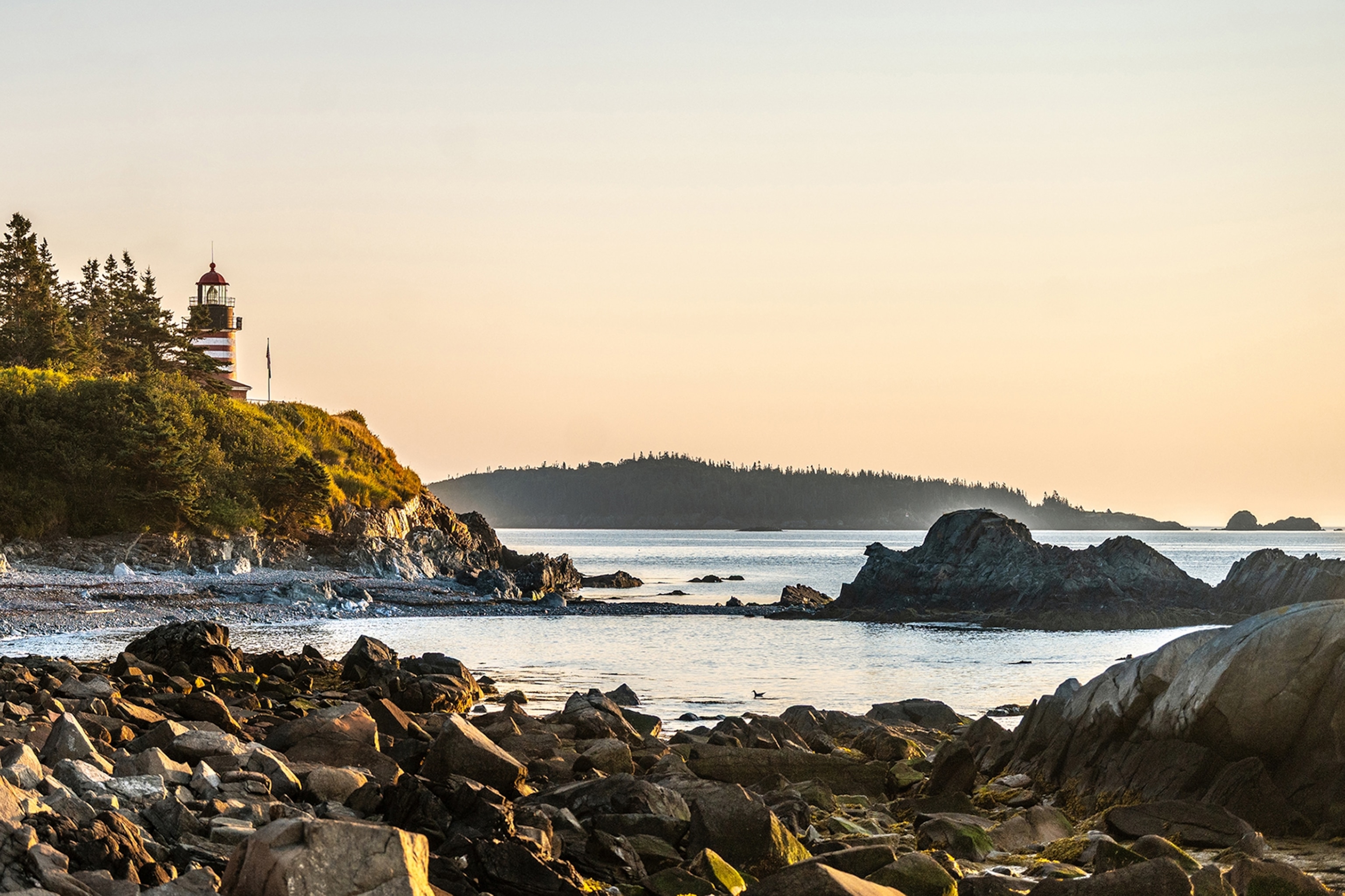 The sunrises out of frame. In the foreground are rocks leading into water, in the middle ground is a small hill to the left with a red and white lighthouse sitting on it, and in the background a hill with trees can be seen in the distance. The sunrise is creating a warm, orange glow across the rocks, hill, and sky in the background.