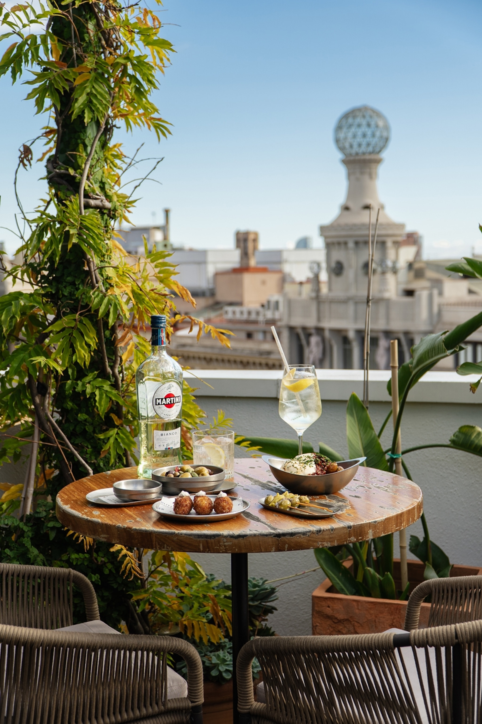 A rooftop terrace with views over Barcelona, focusing on a round table set with wine and food.