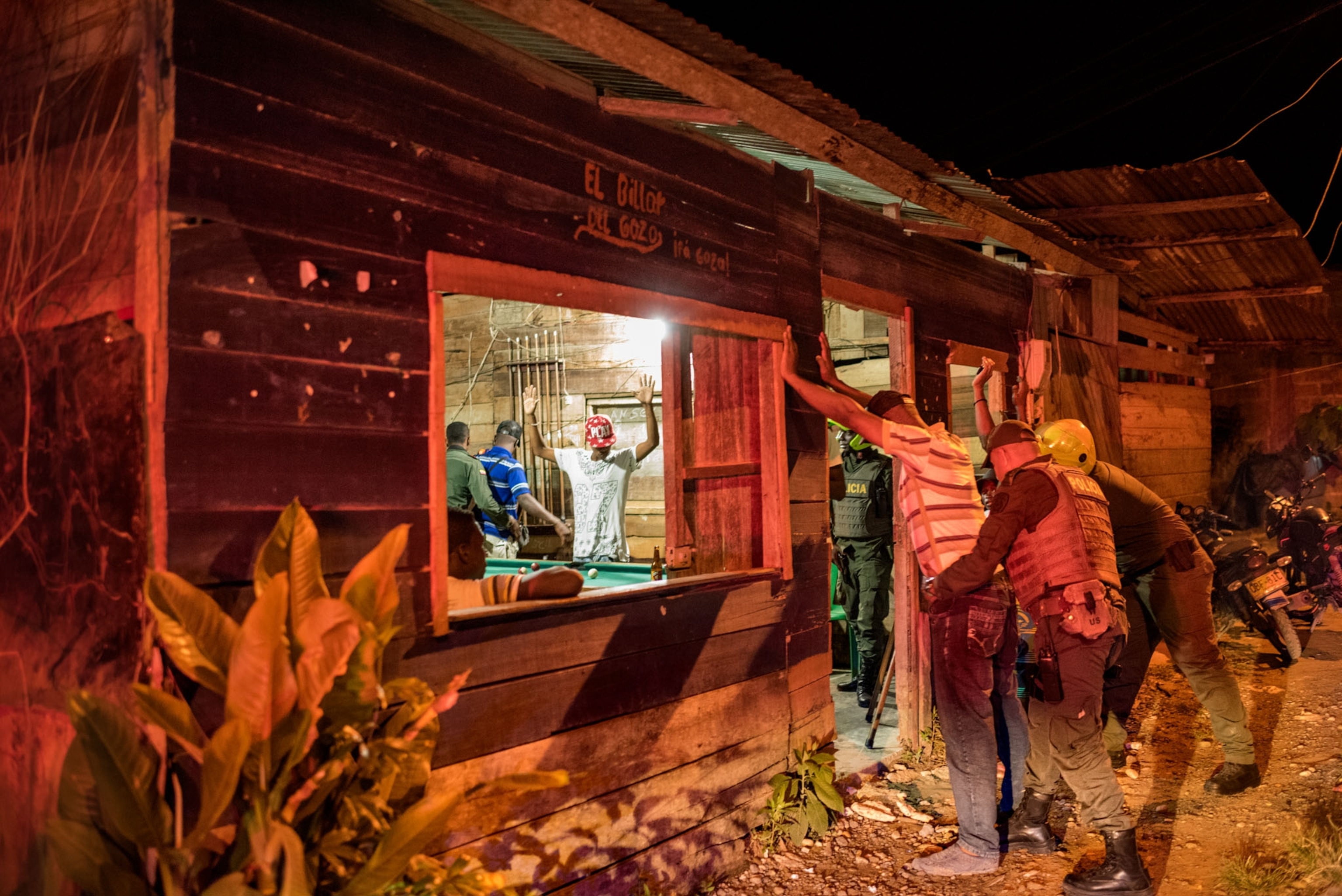 a man beings frisked outside of a bar, inside we can see another man with his arms raised