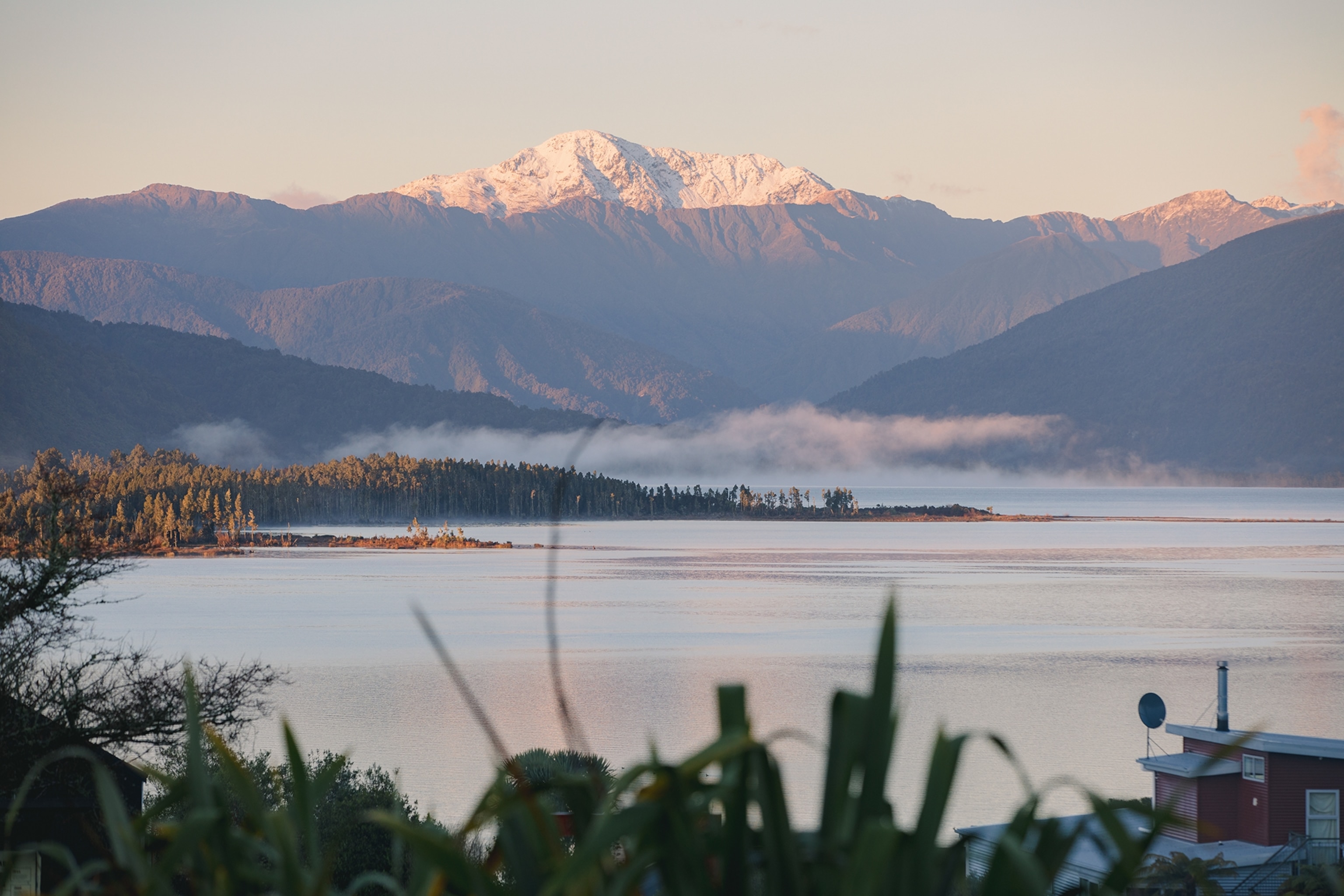 An impressive, wide shot of a calm lake at dawn, line with a pine tree forest and ringed by snow-capped mountains in the distance.