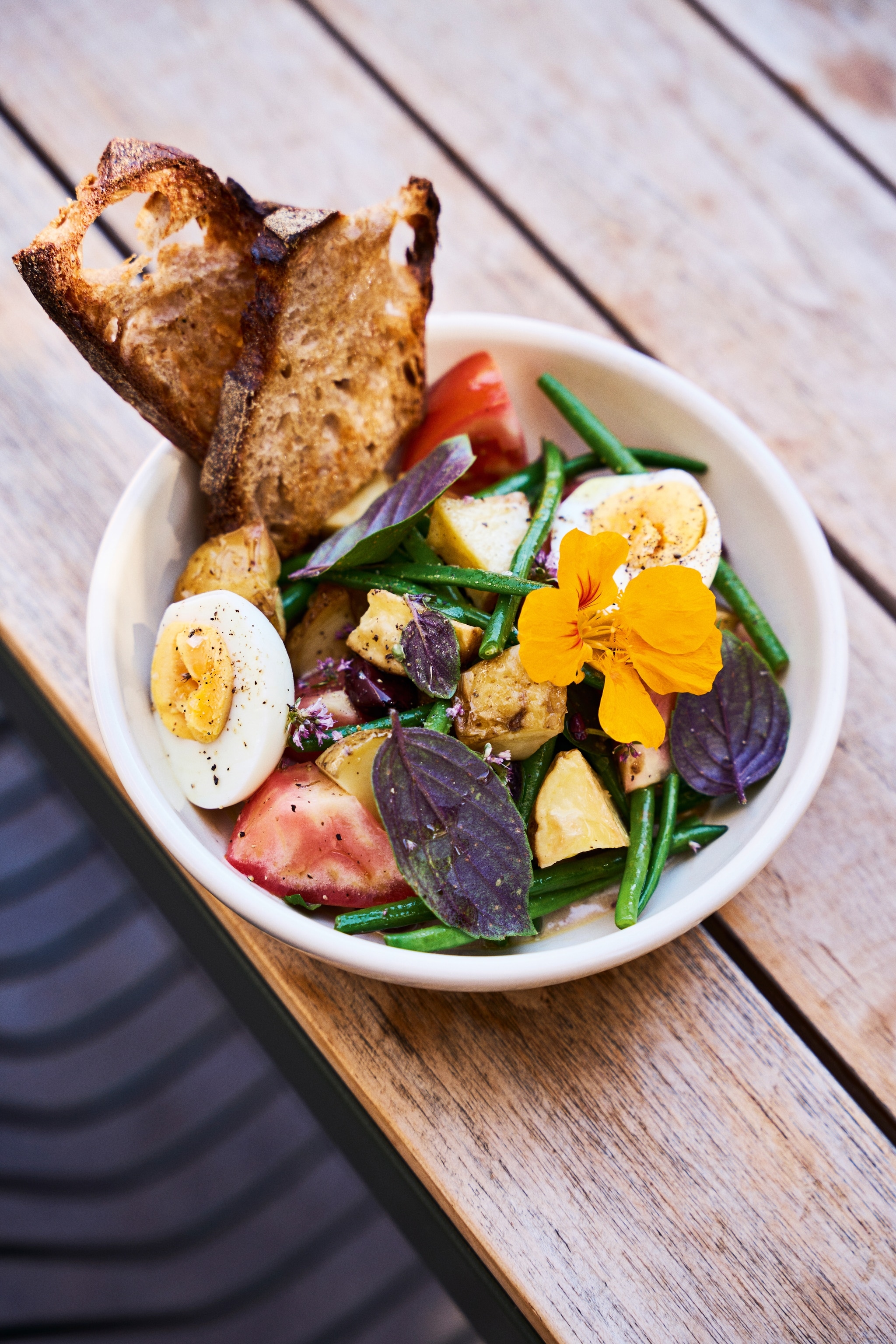 A bowl of sauteed green beans topped with hard boiled eggs and served with two toasts on an outside patio table.