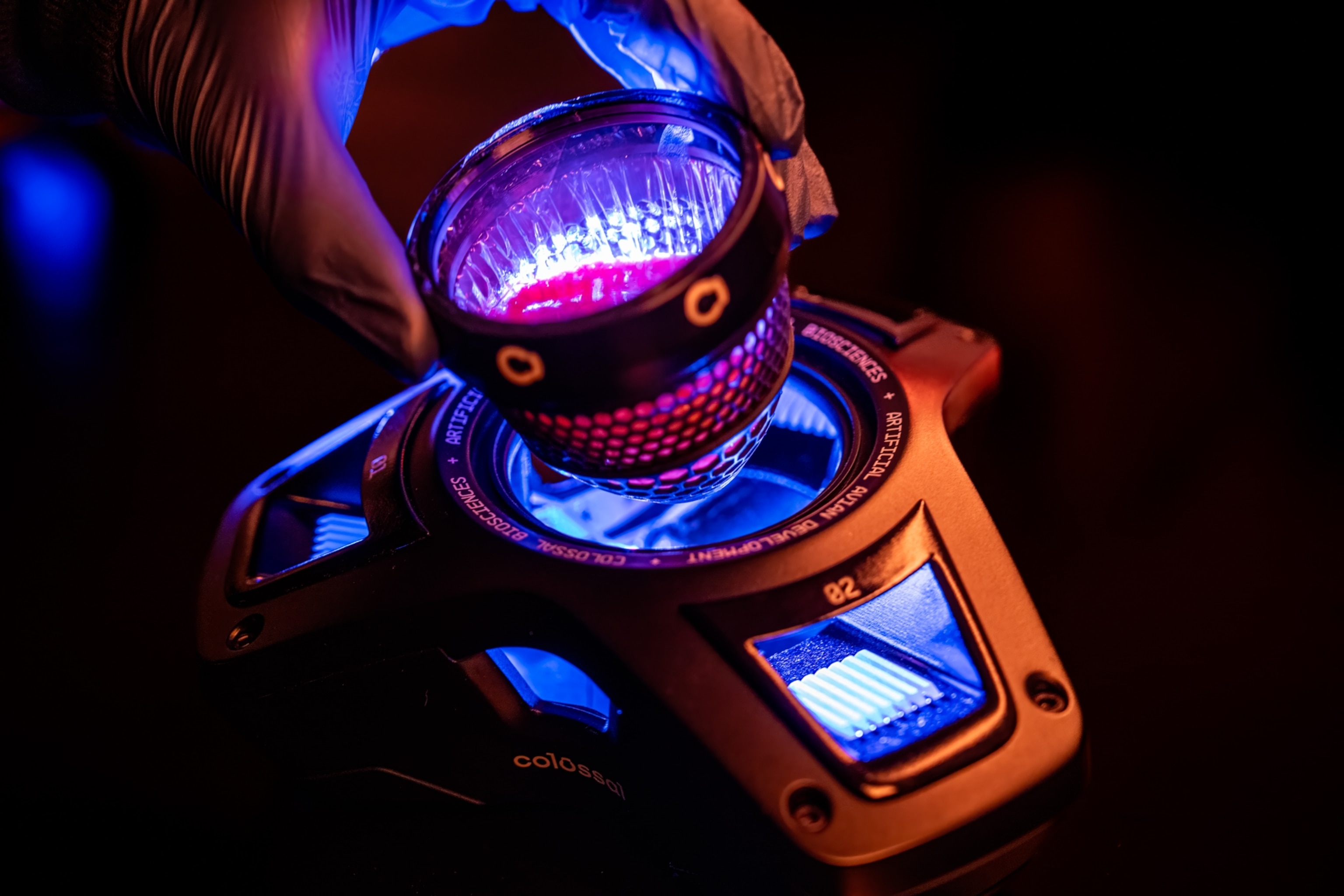 A hand in a glove holding a basket-like cup over a futuristic device emitting a bright blue and red glow