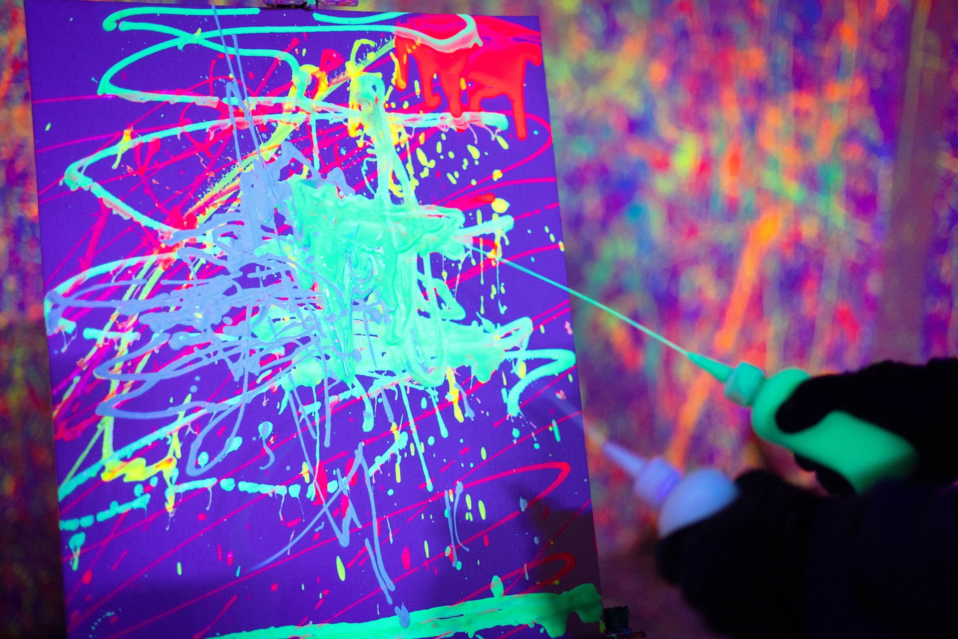 An easel in a rage room with someone squirting bioluminescent colour onto the canvas.