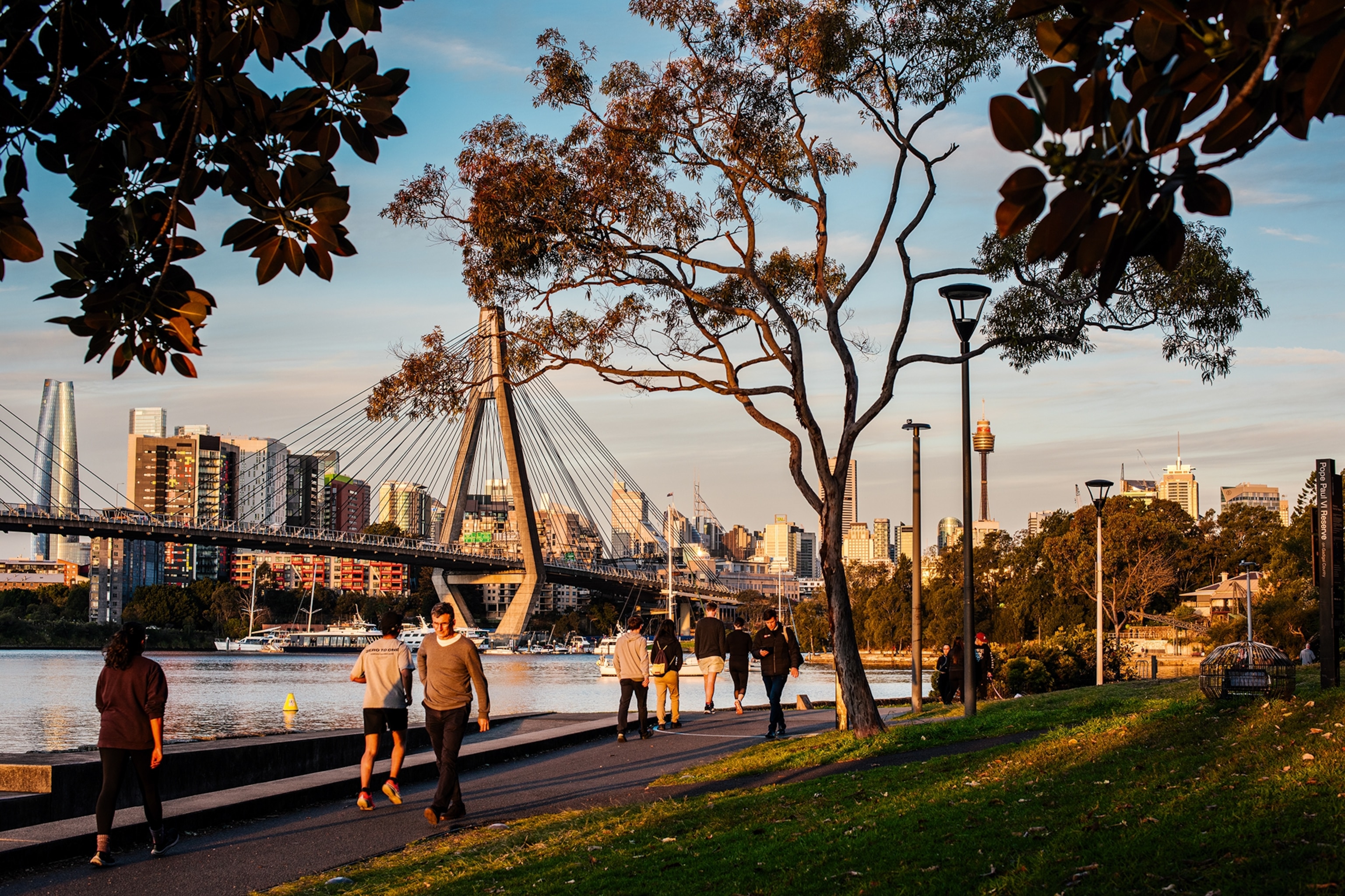 An urban river promenade at sunset with a bridge connecting both sides of the river and the skyline in the background.