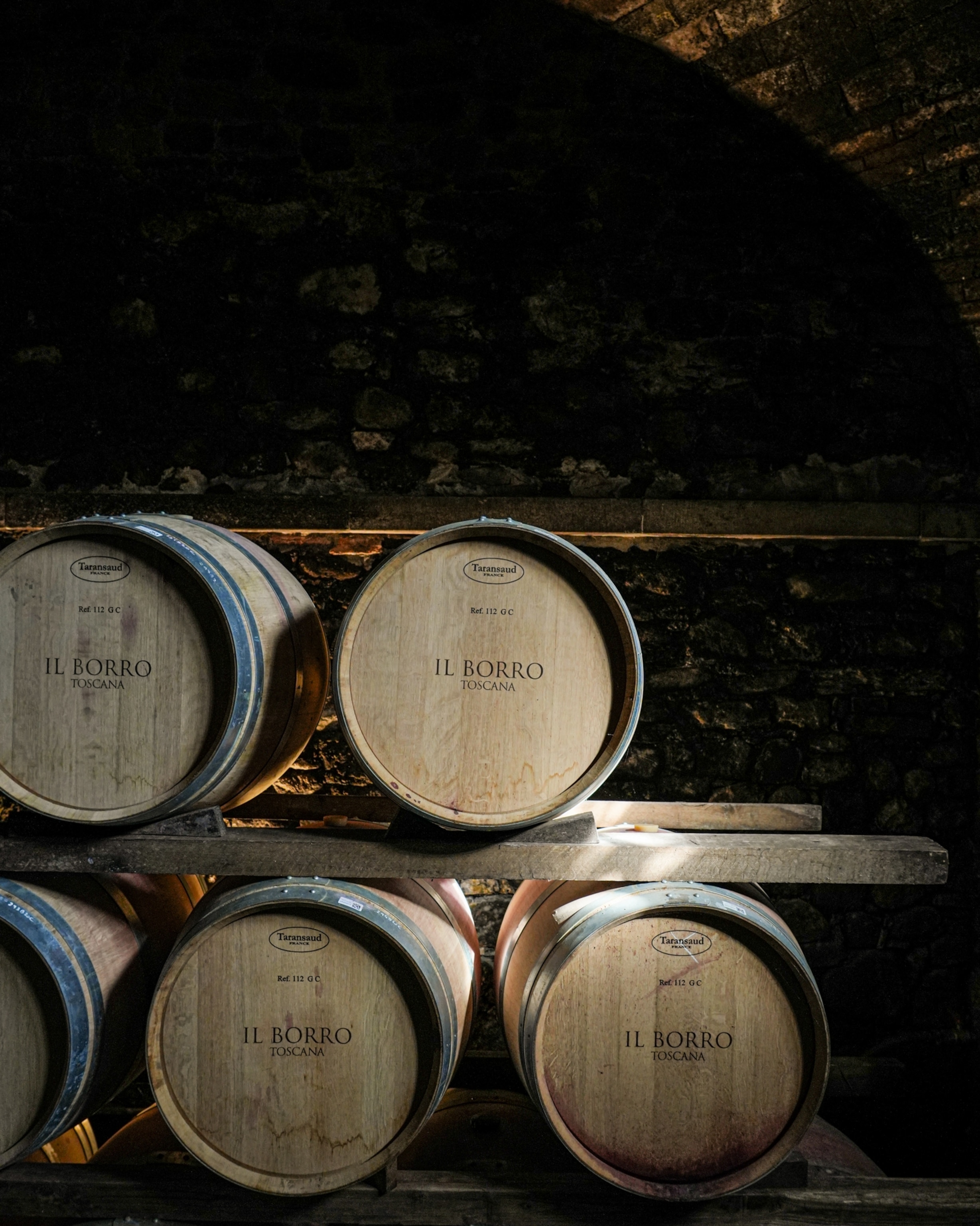Multiple wood wine barrels stacked on top of each other in a dark wine cellar at the Il Borro Toscana hotel.