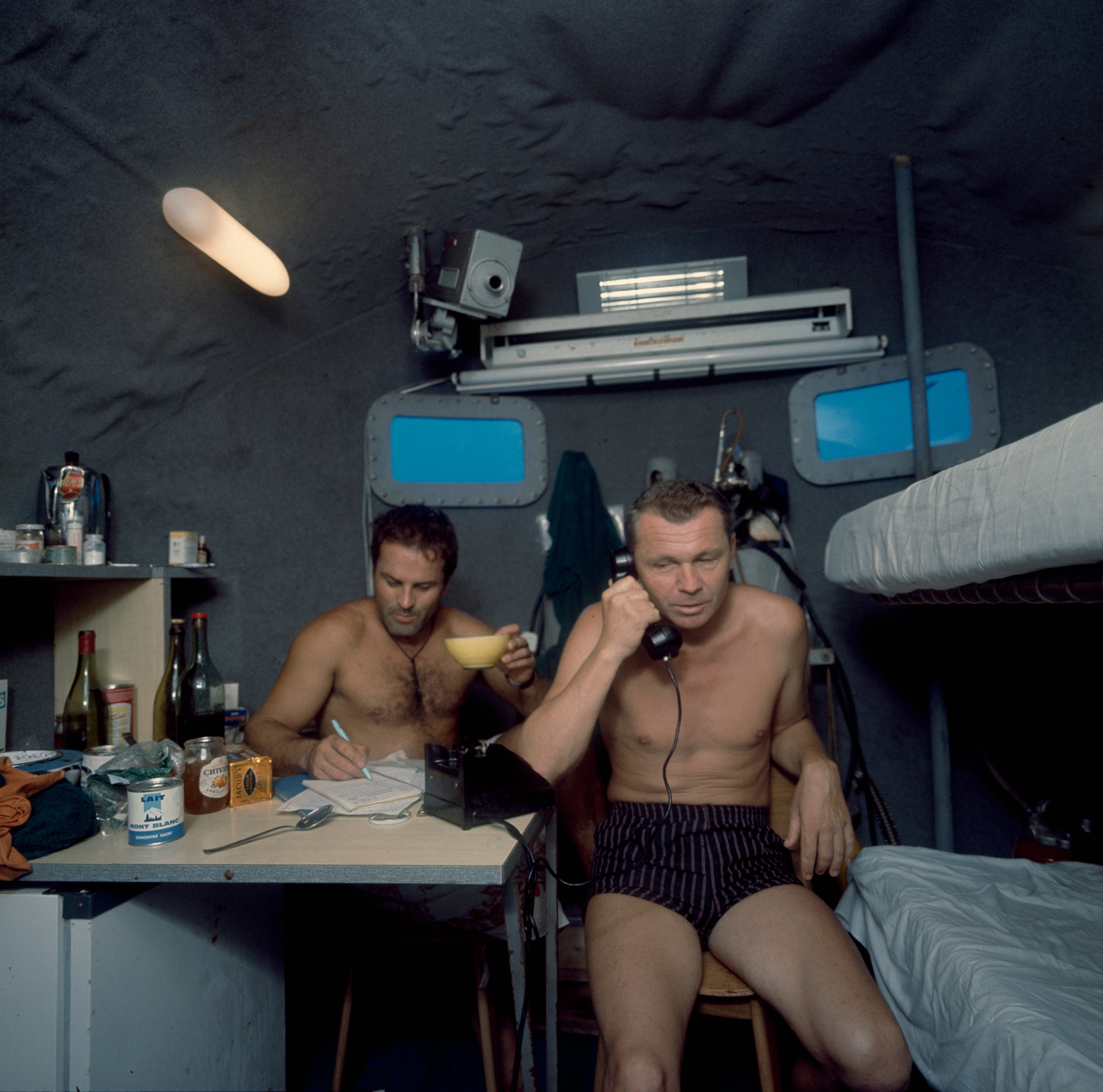 Men living in a submersible talk to colleagues on a telephone.