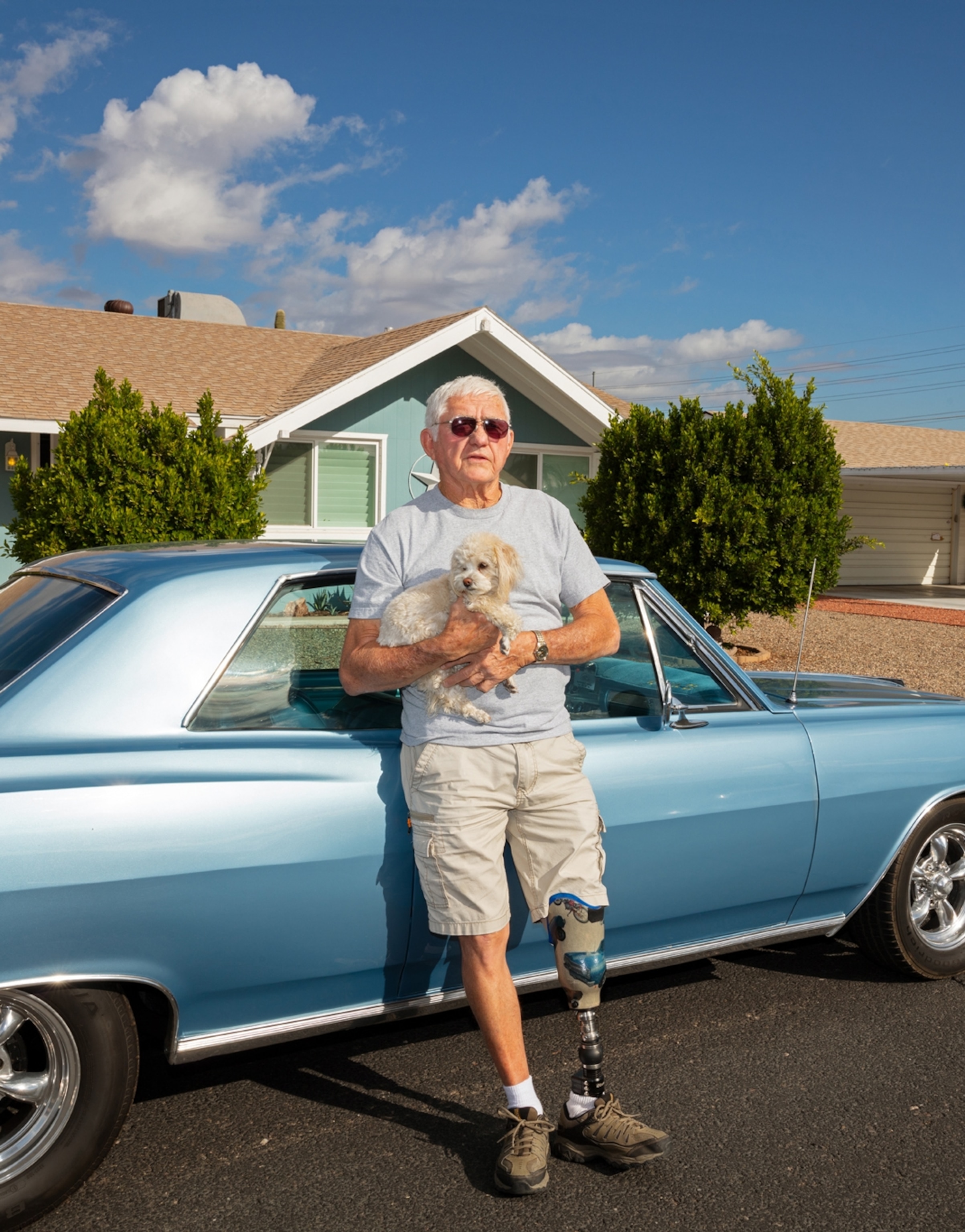 Picture of man with prosthetic leg holding a small dog next to the vintage car.