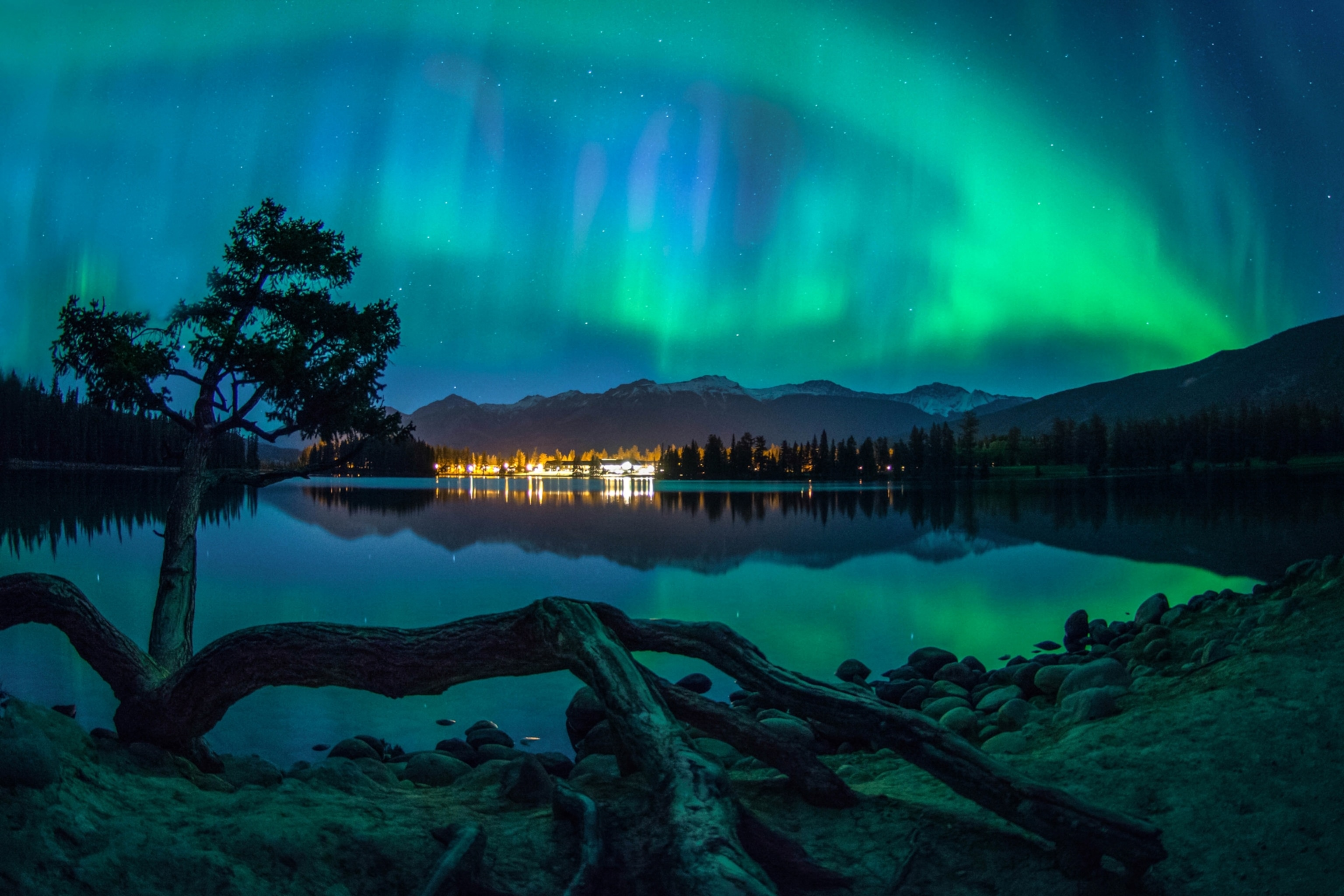 Green and blue northern lights glow across the night sky above a calm lake, with mountains and trees reflected in the water.