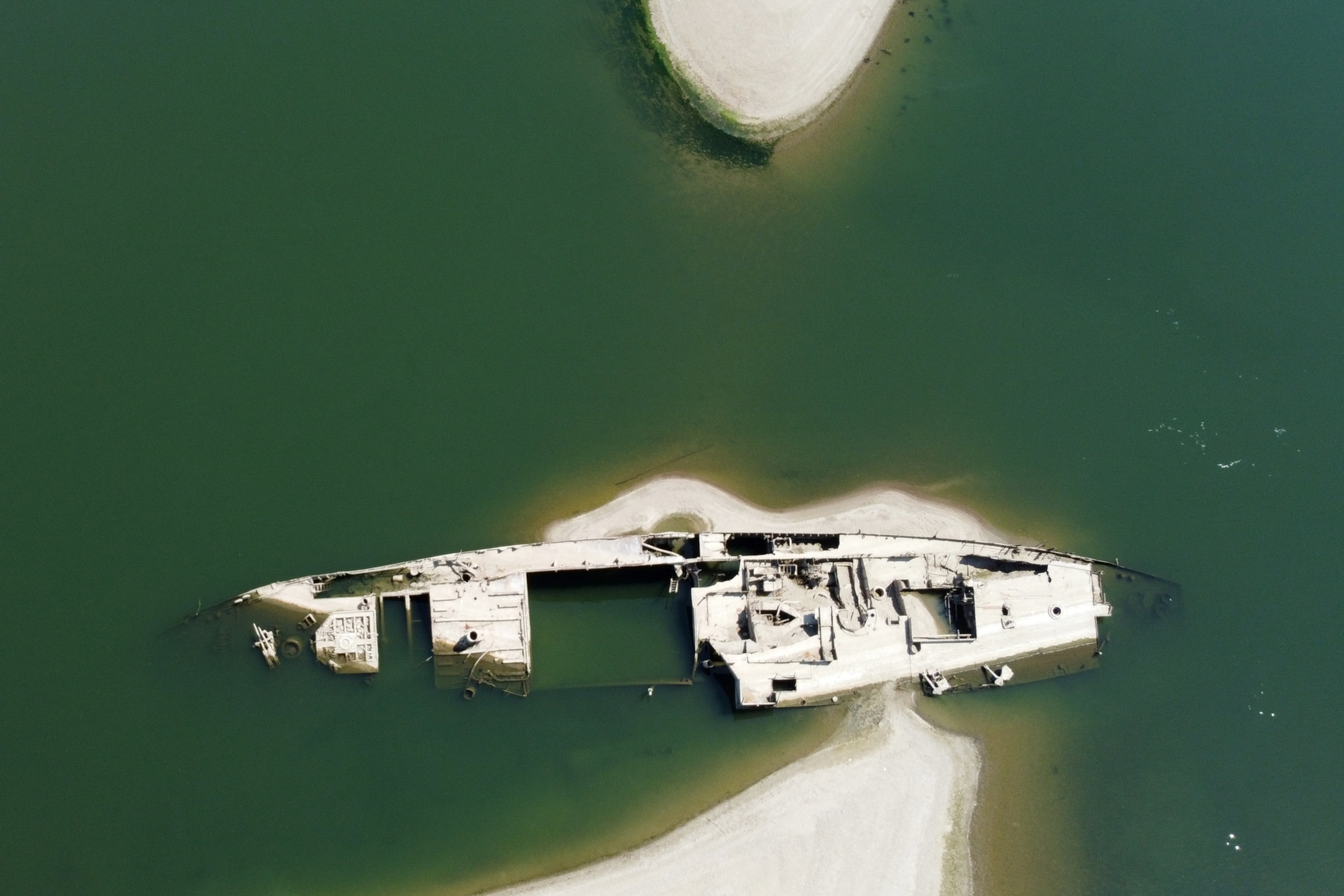 An aerial view of a rusted, partially submerged shipwreck resting on a sandy riverbank surrounded by deep green water
