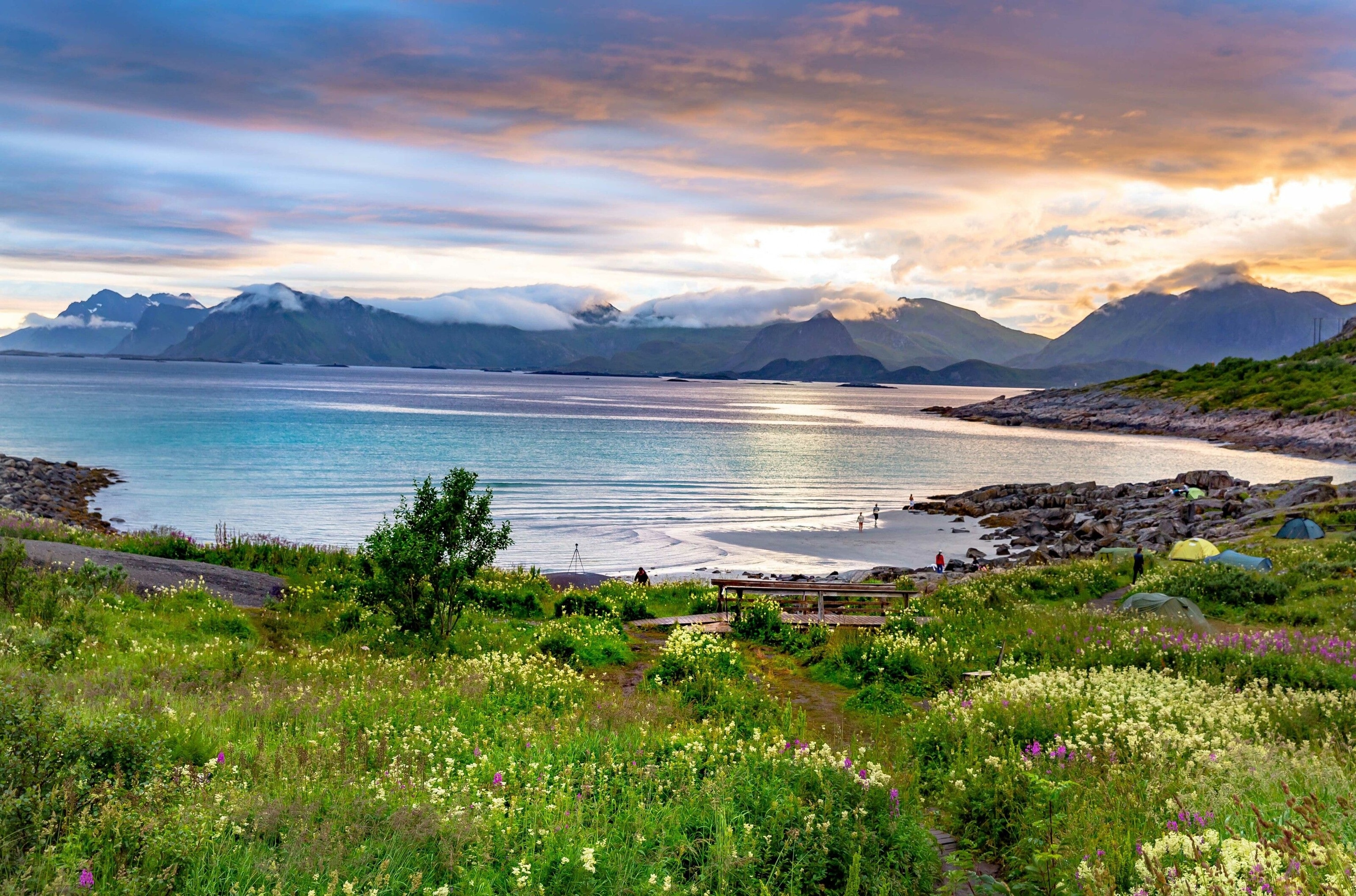 Lush green fields spill into calm blue water. Mountains surround.