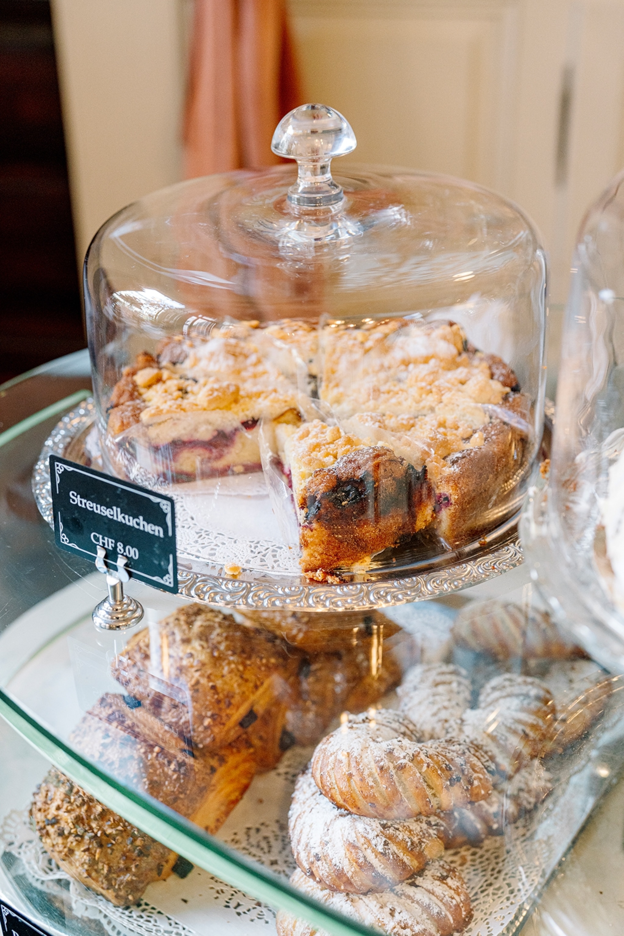 A streusel - crumble - cake in a luxurious cake display with more pastries presented on a glass shelf below.