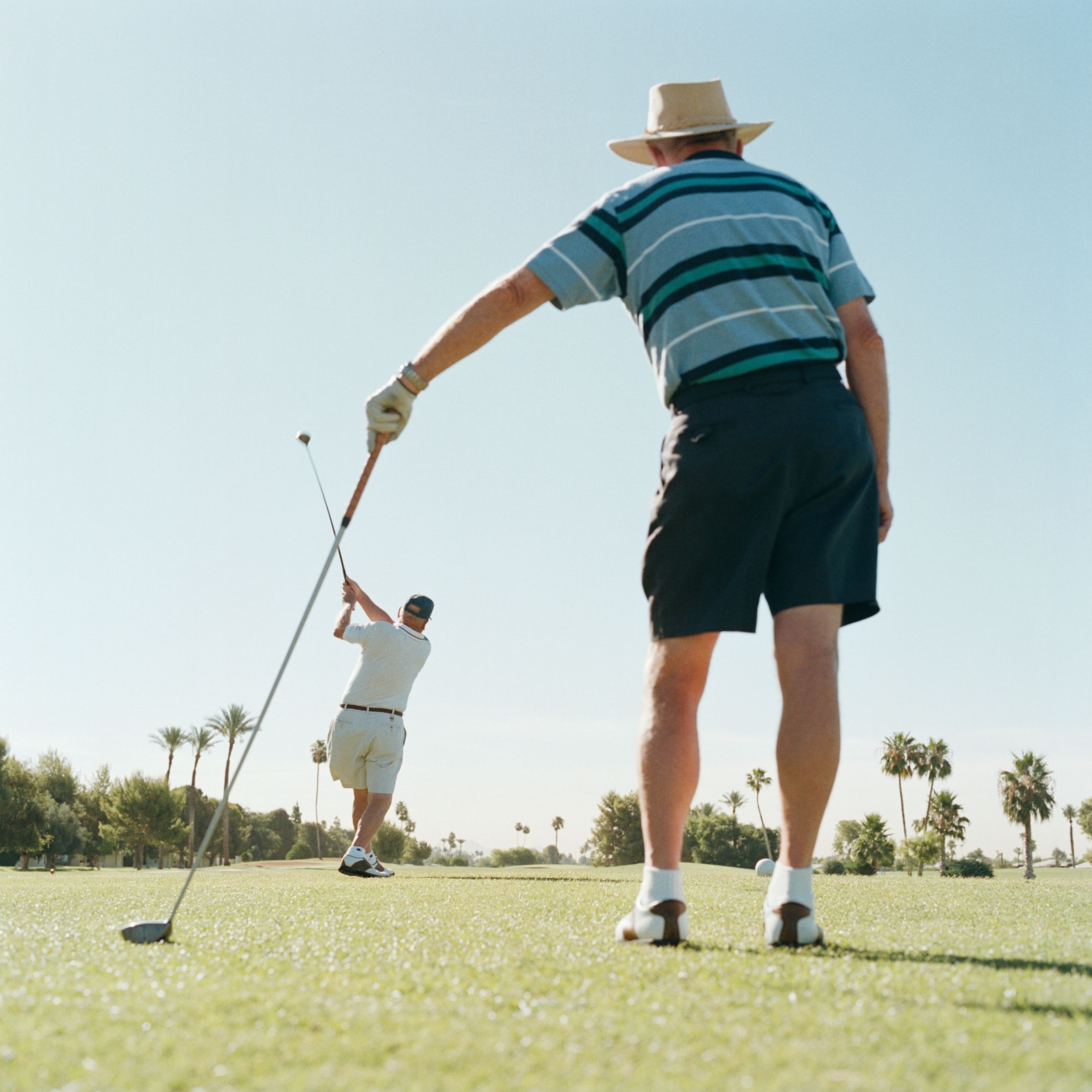 A golfer mid-swing, seen from behind framed between another golfer in the foreground and a golf club he is using to help support his weight