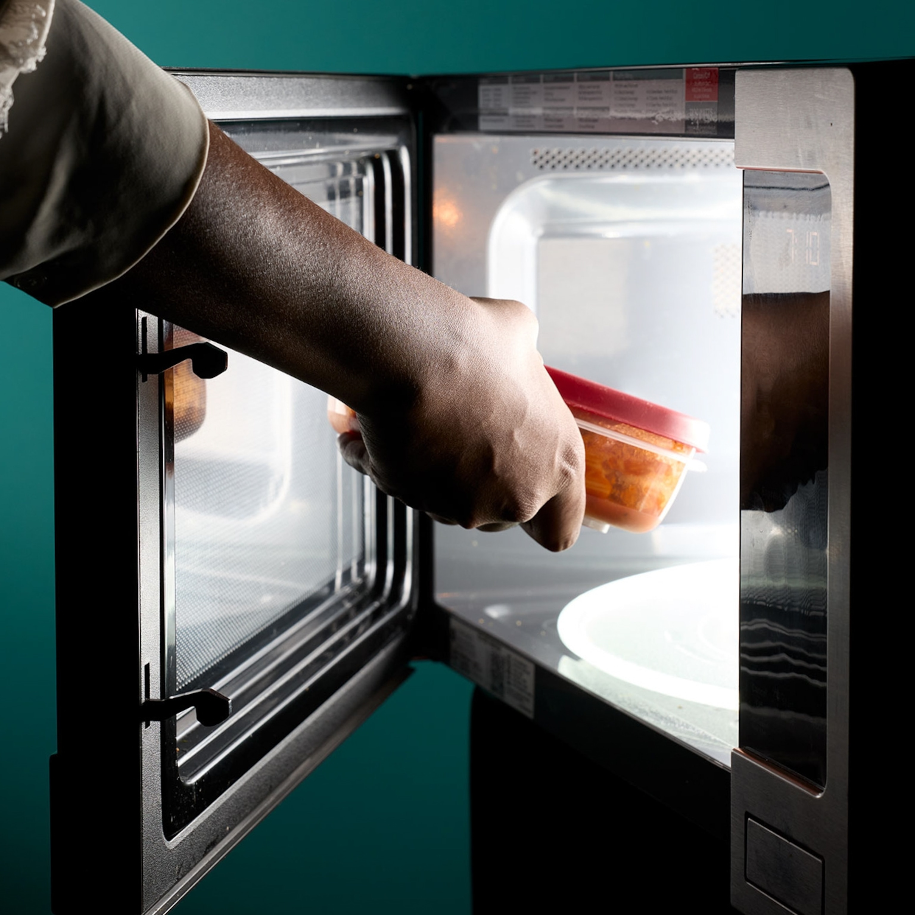 A hand with dark skin tone places a container in a microwave with glowing light.