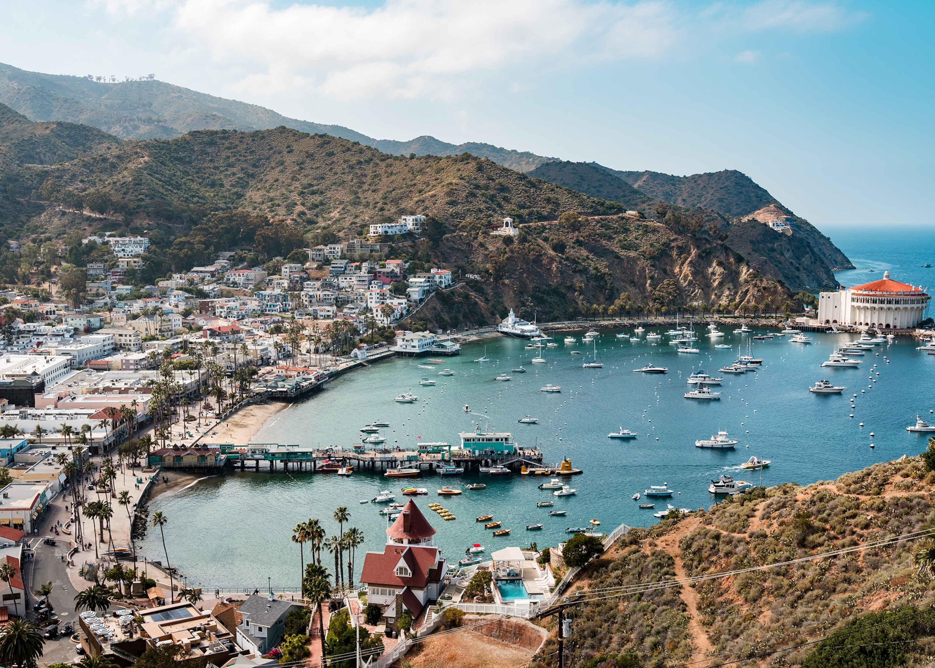 An aerial view of a coastal harbor town shows boats anchored in turquoise water, colorful buildings lining the waterfront, and rugged hills rising above the bay under a partly cloudy sky.