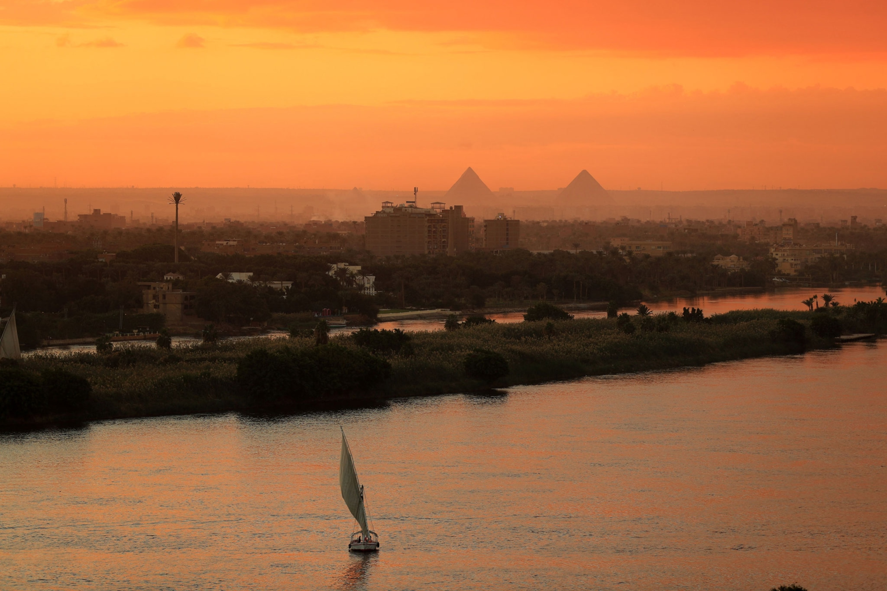 shows a view of the Nile River at sunset in Cairo, Egypt with a sail boat in the foreground and pyramids in the background.