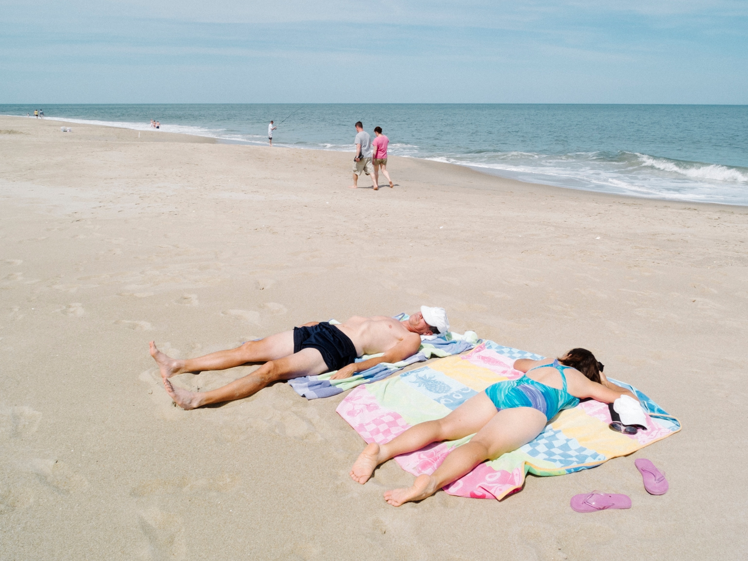 a man and a woman lying on pastel beach towels on a sandy beach as people walk by in the background