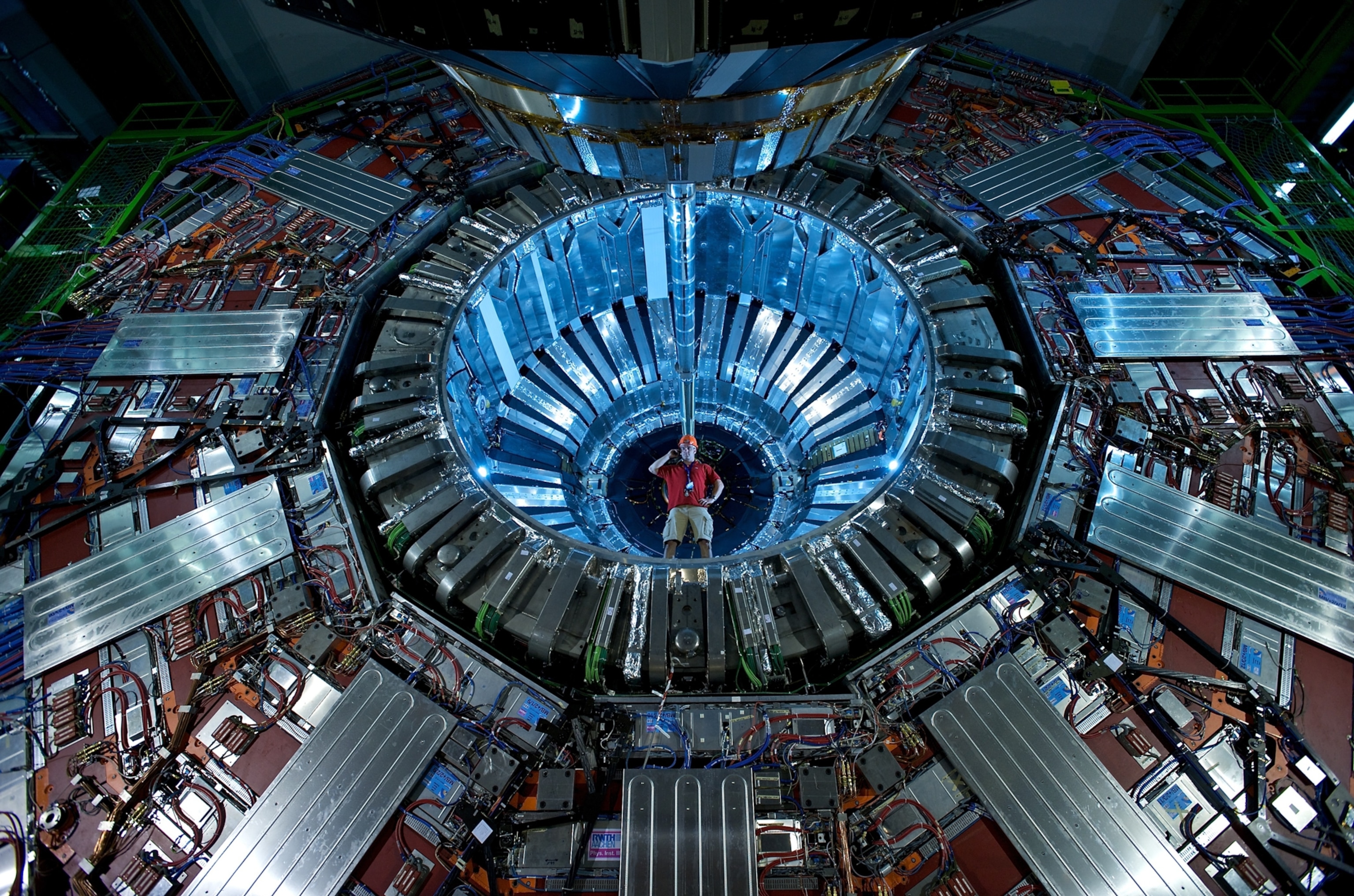 A researcher wearing a hardhat stands at the center of a large, circular particle detector, surrounded by intricate layers of metal and wiring. The scene is illuminated with blue light, conveying a high-tech and futuristic atmosphere.