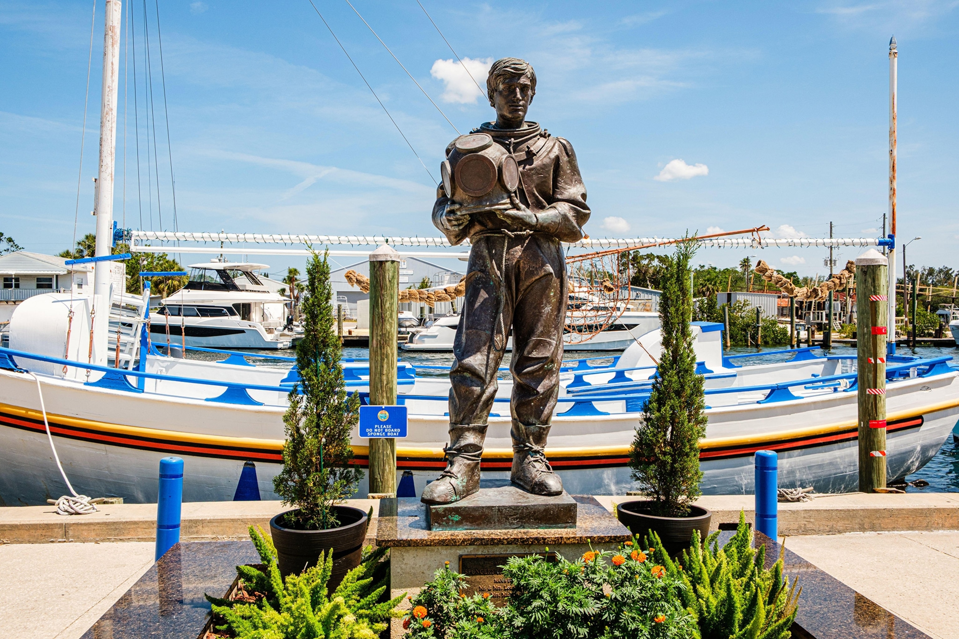 A bronze statue of a diver with a helmet in hand stands on a dock with boats behind