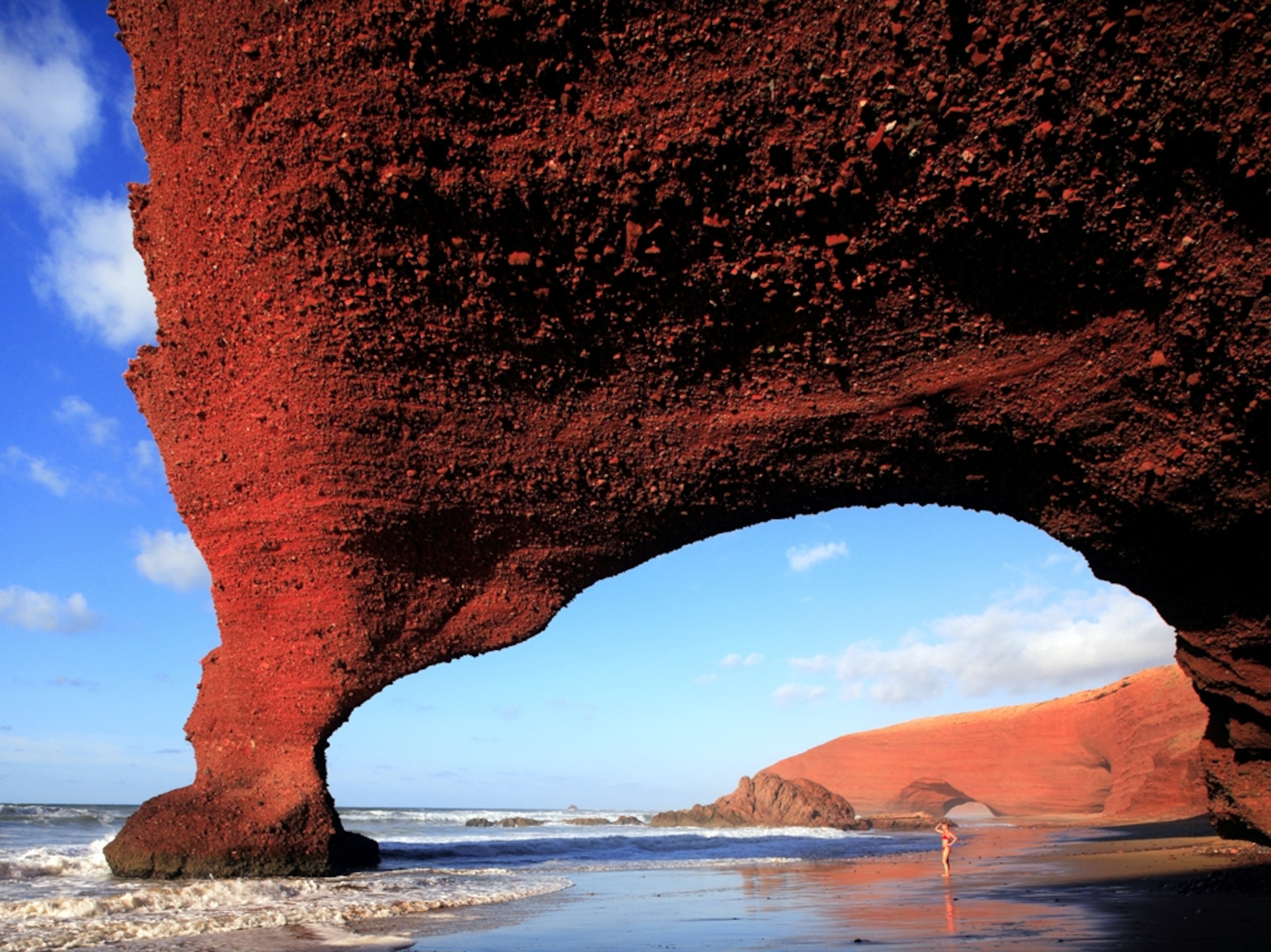 a woman standing under a large arch at Legzira Beach, Morocco