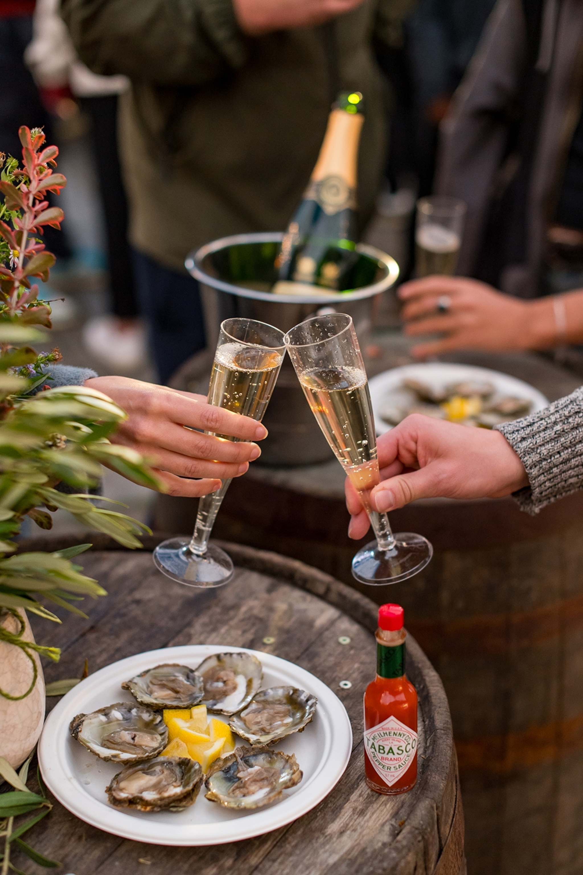 A plate of oysters on a barrel next to a bottle of Tabasco sauce as two people cheers prosecco glasses.