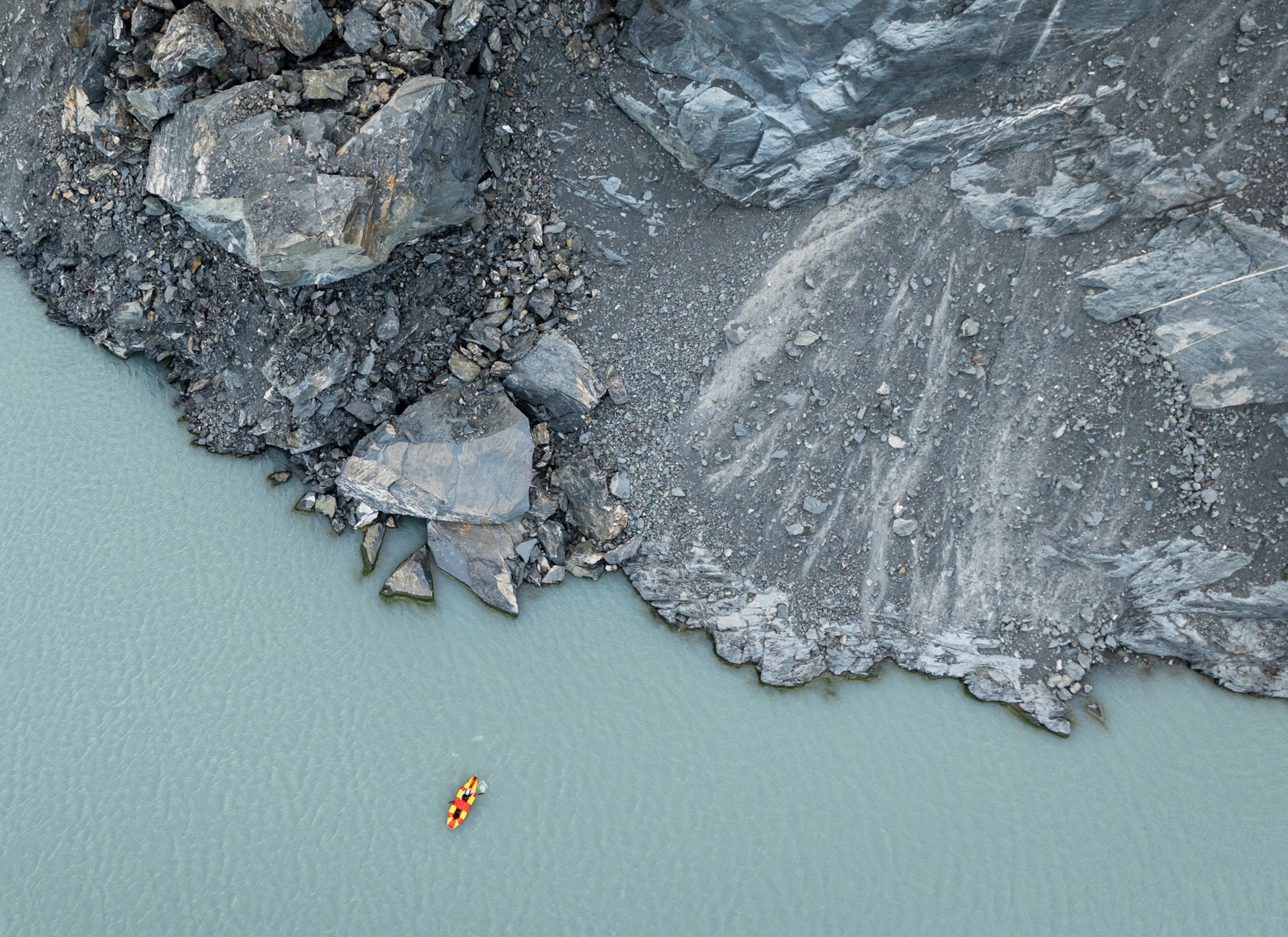 A view from above shows gray rocks and stones next to light blue water. The rocks are sharp and broken along the shore. In the water, near the bottom of the image, a small red and yellow kayak floats by itself.