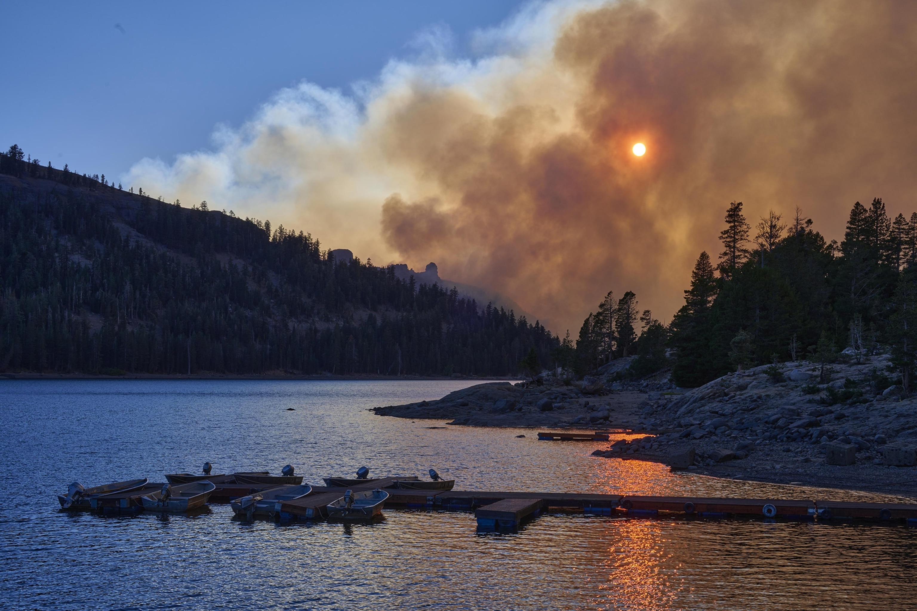 Thick smoke obscures the sun above a mountain and lake landscape, a pier with boats in the foreground.