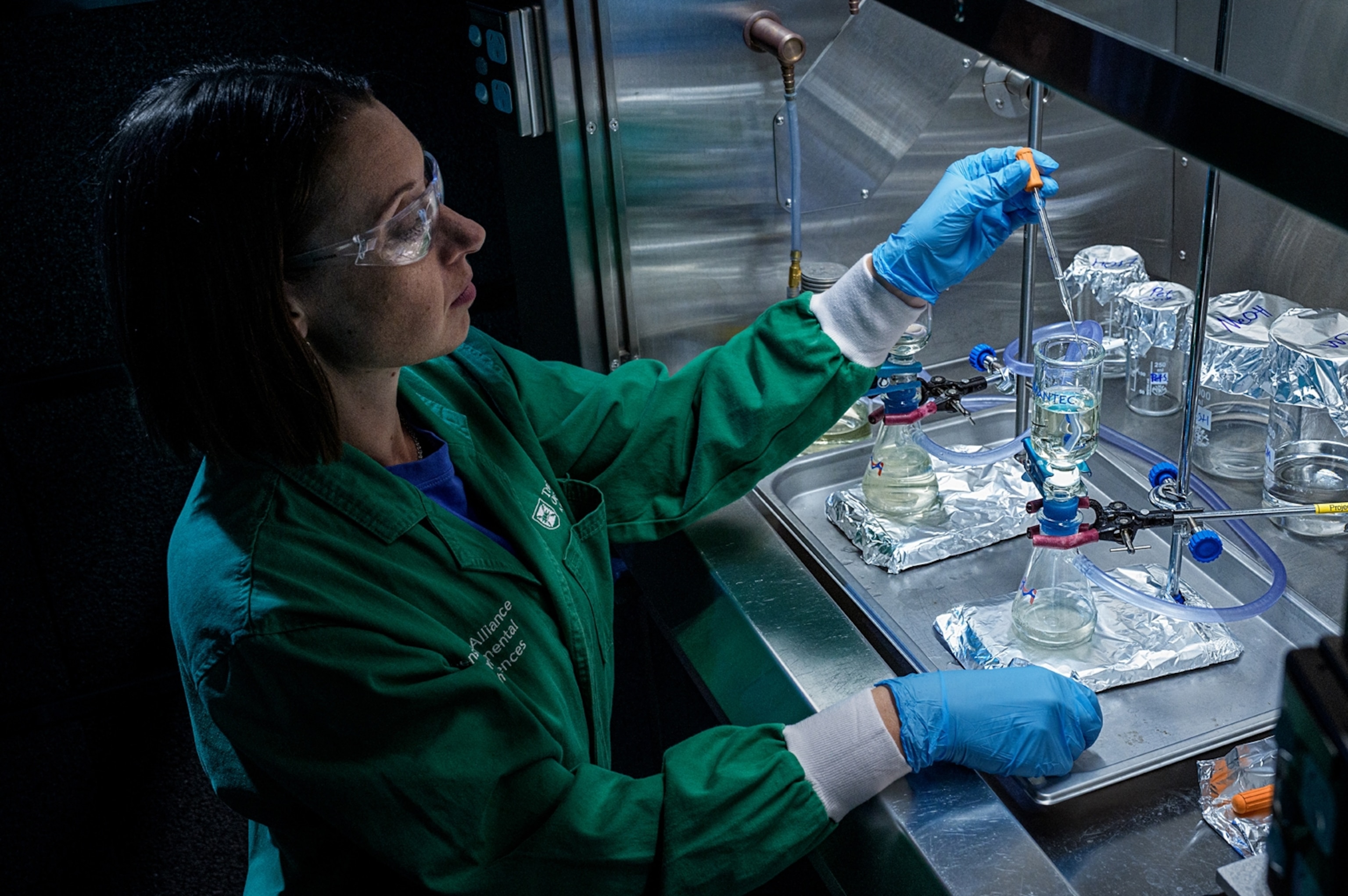 A woman in scientific protective gear works with a dropper and solution and beakers in a lab