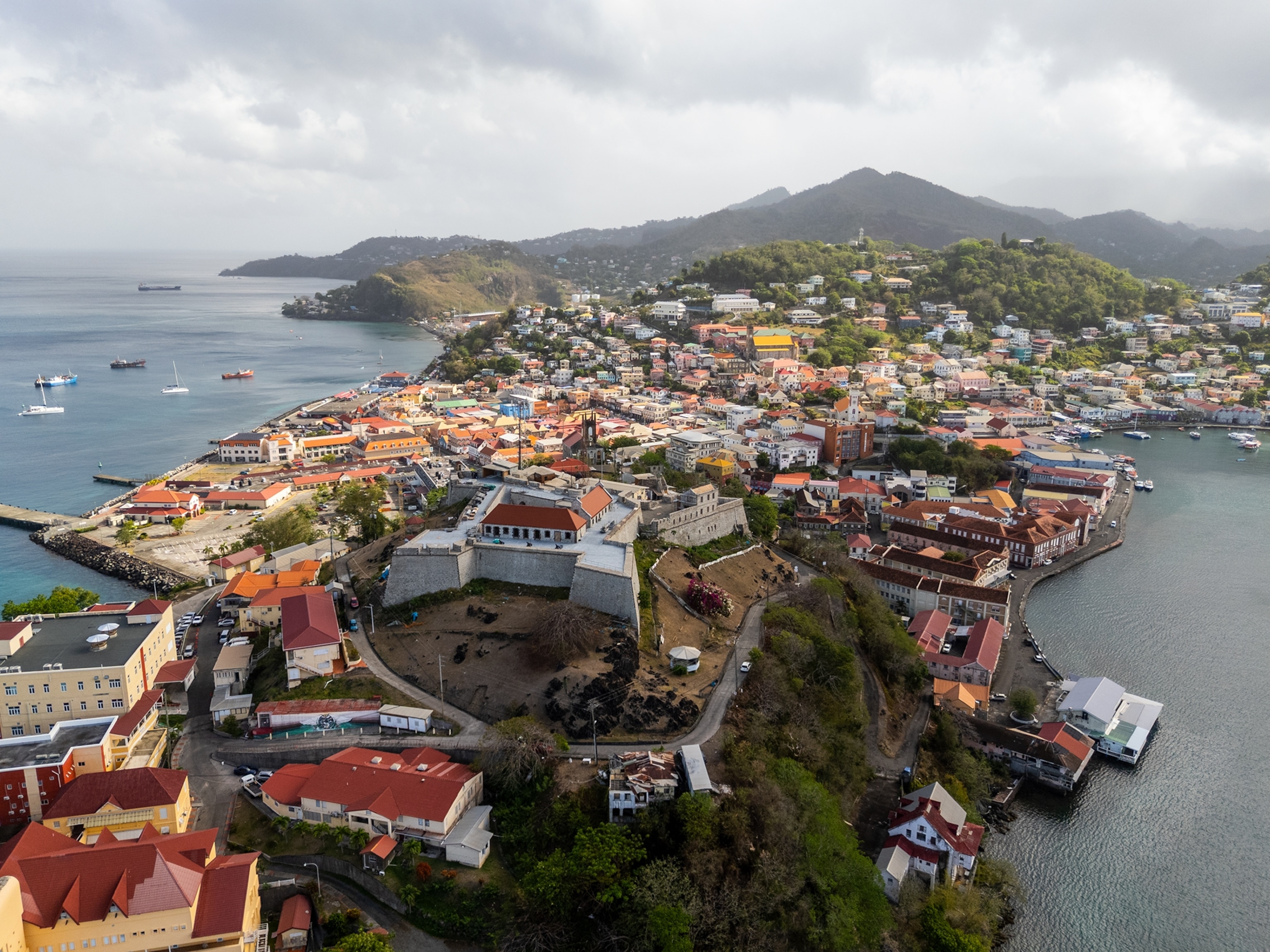 An aerial view of a coastal city built around a harbor, with colorful buildings, a historic hilltop fort, boats along the waterfront, and green mountains rising in the background under cloudy skies. 