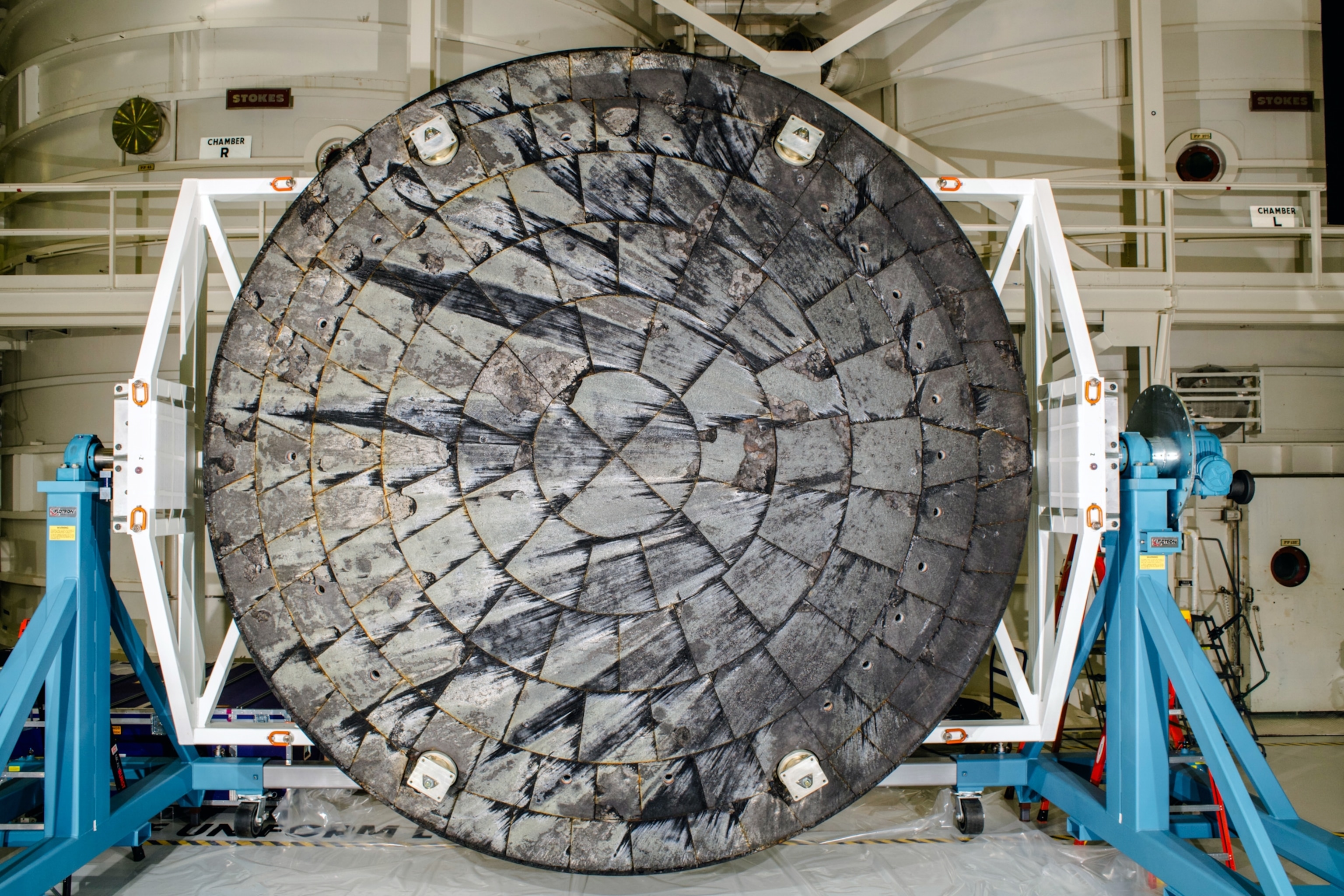 A circular, heat-scorched spacecraft heat shield displayed vertically, surrounded by a sturdy white frame and blue support stands in an industrial setting.