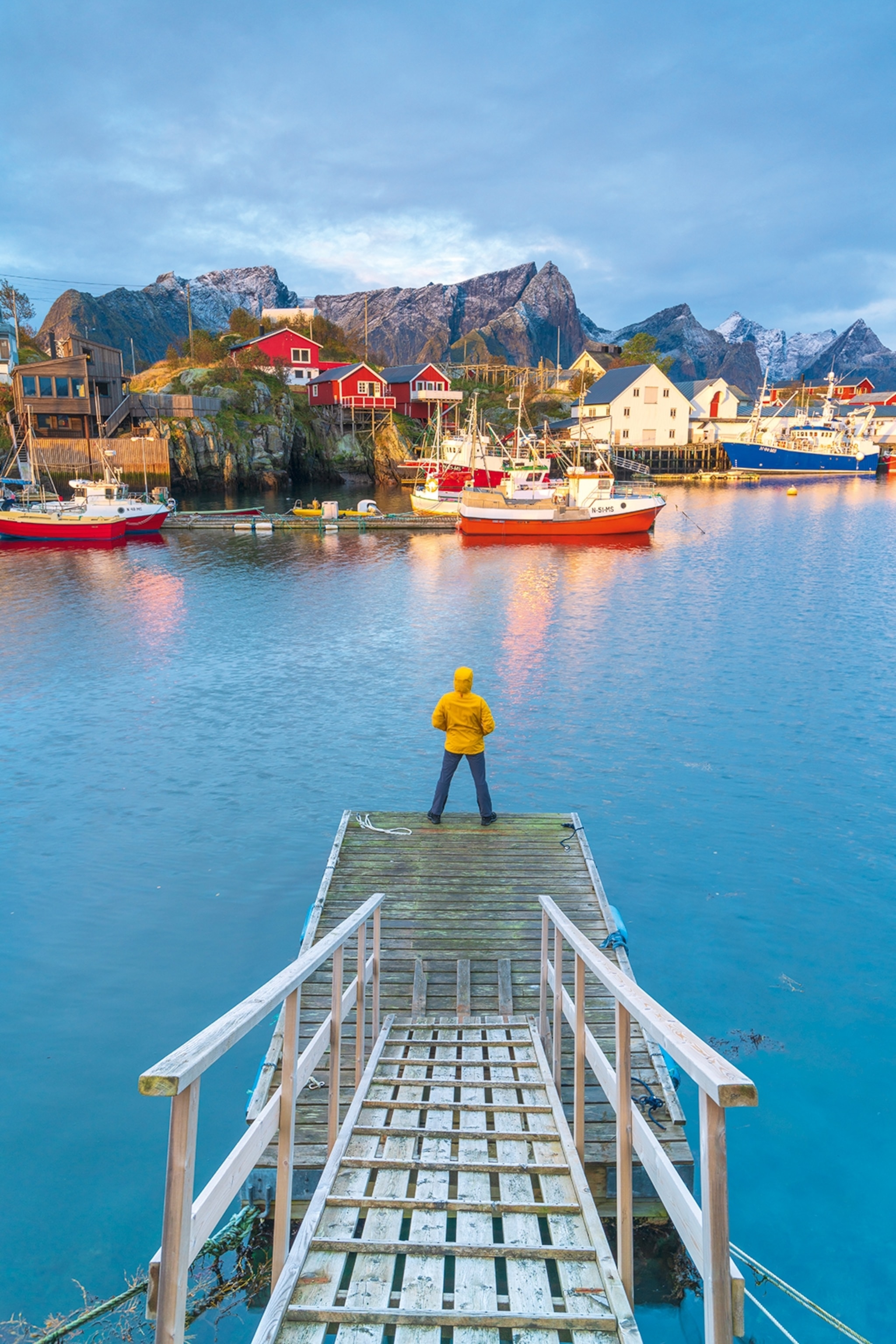 A person standing at the far end of a pier in a fjord, looking at a small harbour town in Norway.
