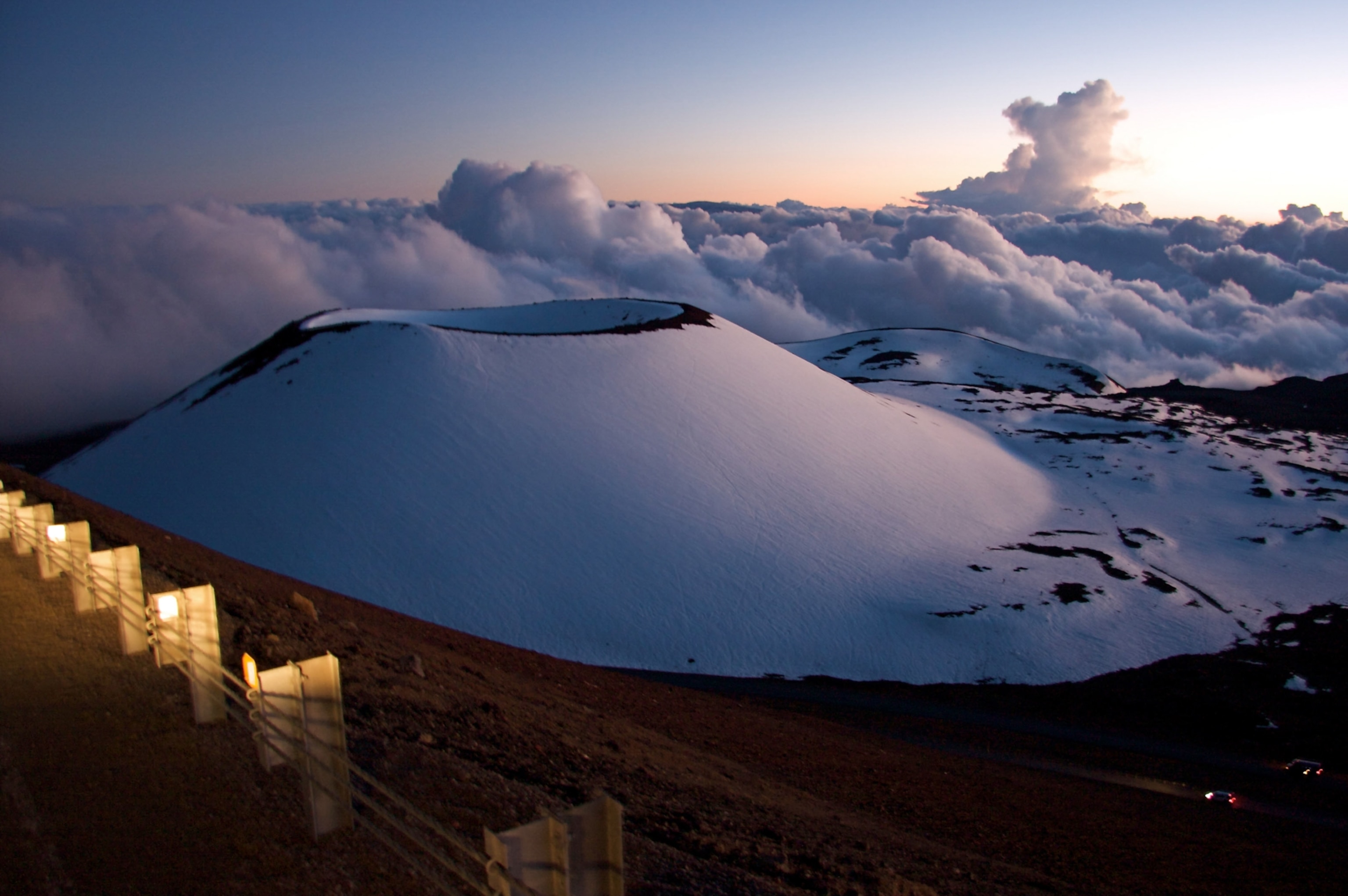Snow-covered volcanic peaks rise above a sea of clouds at sunset, with a glowing horizon and a trail of lights lining the path in the foregrou
