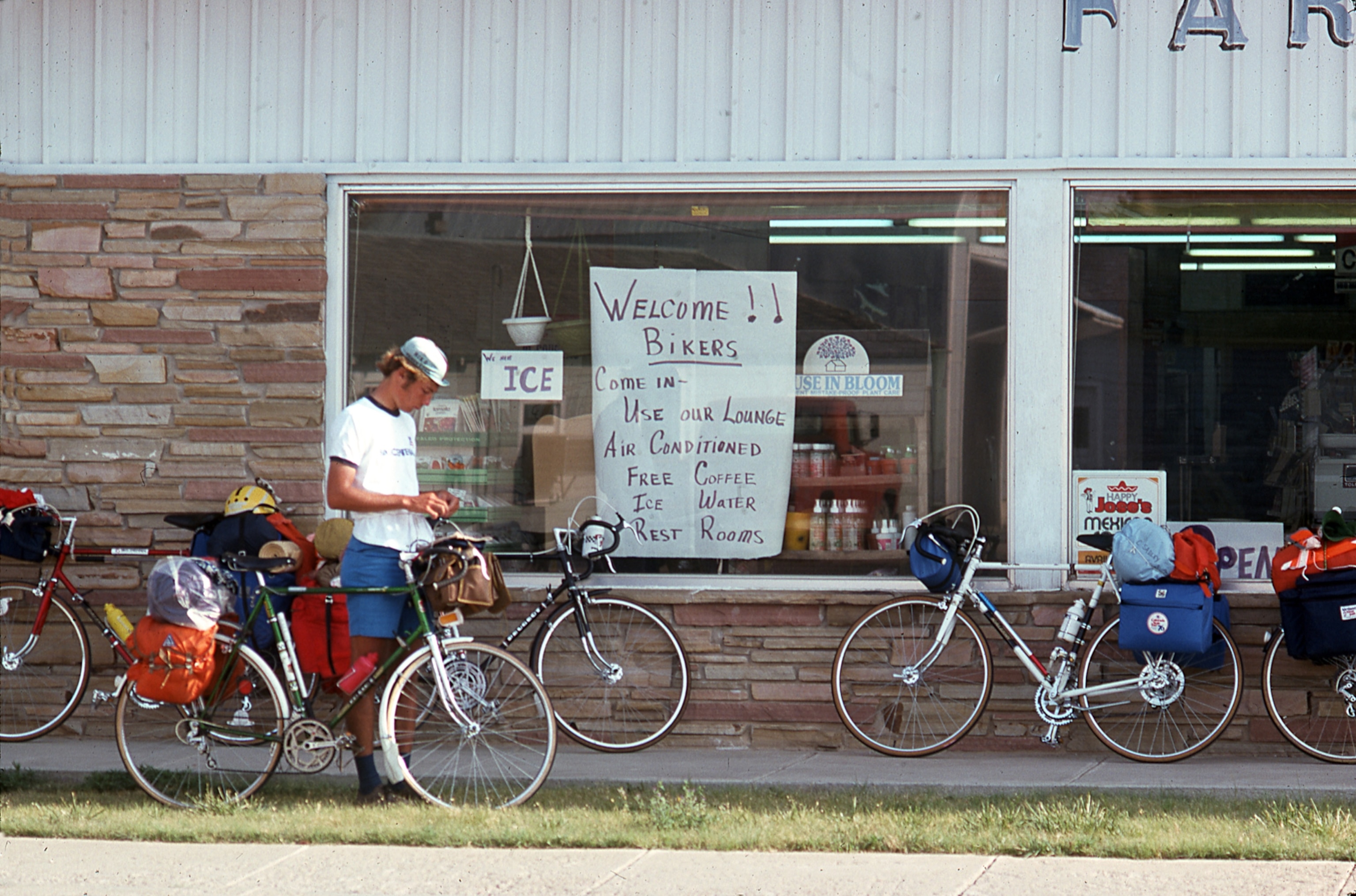 A man wearing a white ti shirt and a white hat stands behind his bike while taking a rest in front of a grocery store.
