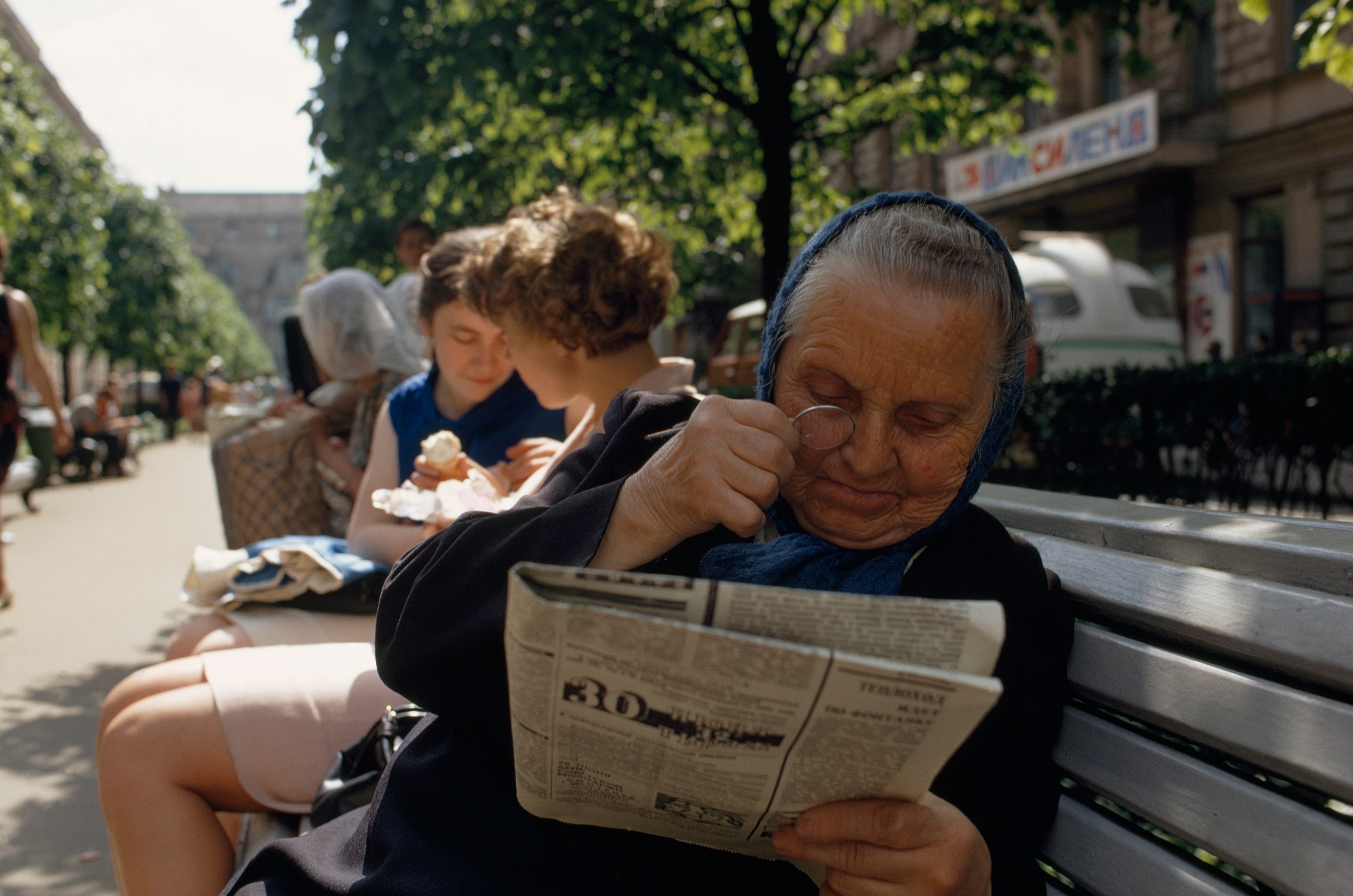 An elderly woman reads a newspaper on a bench using a monocle.