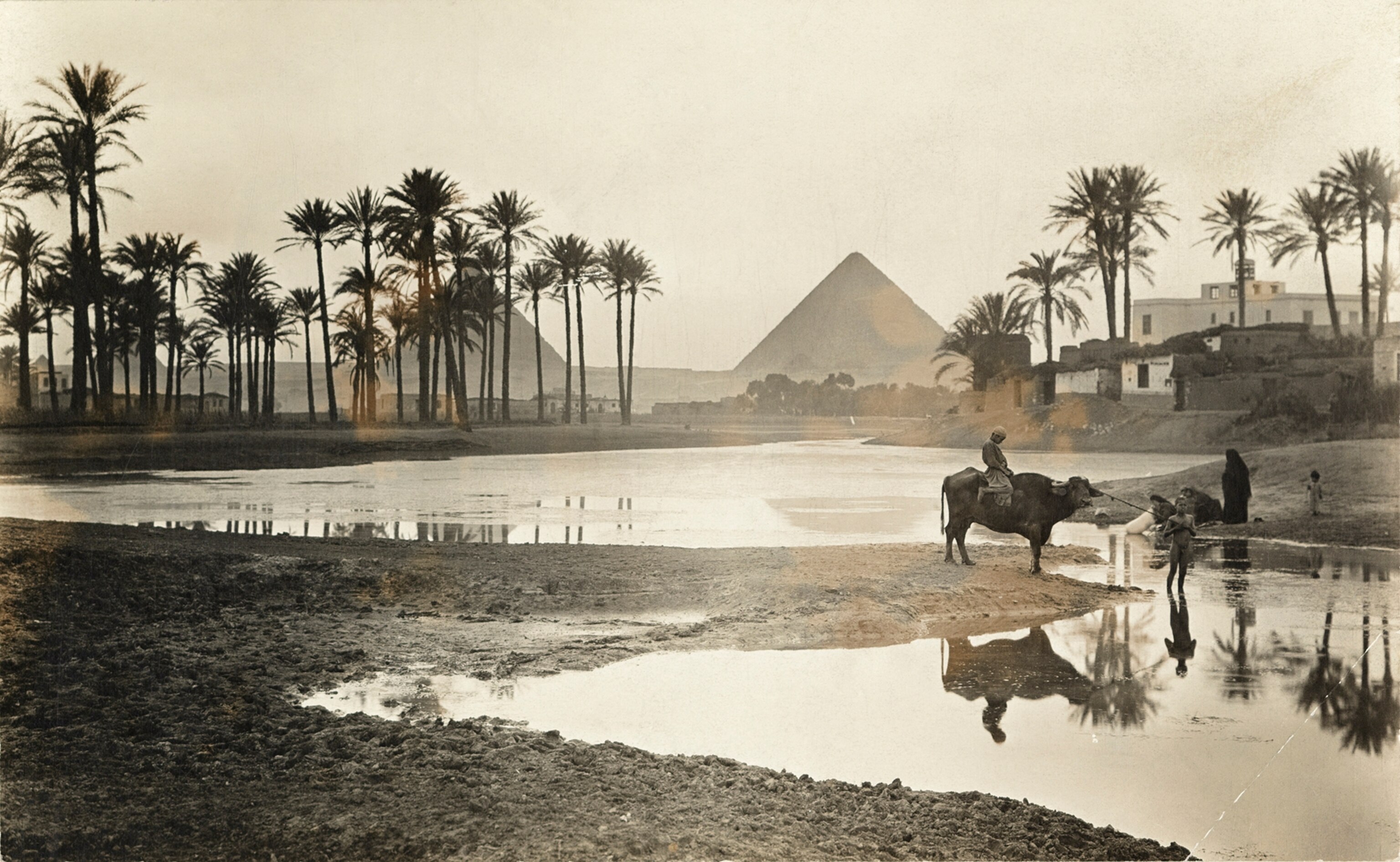 Villagers stand next to water with the pyramids of Giza behind them.