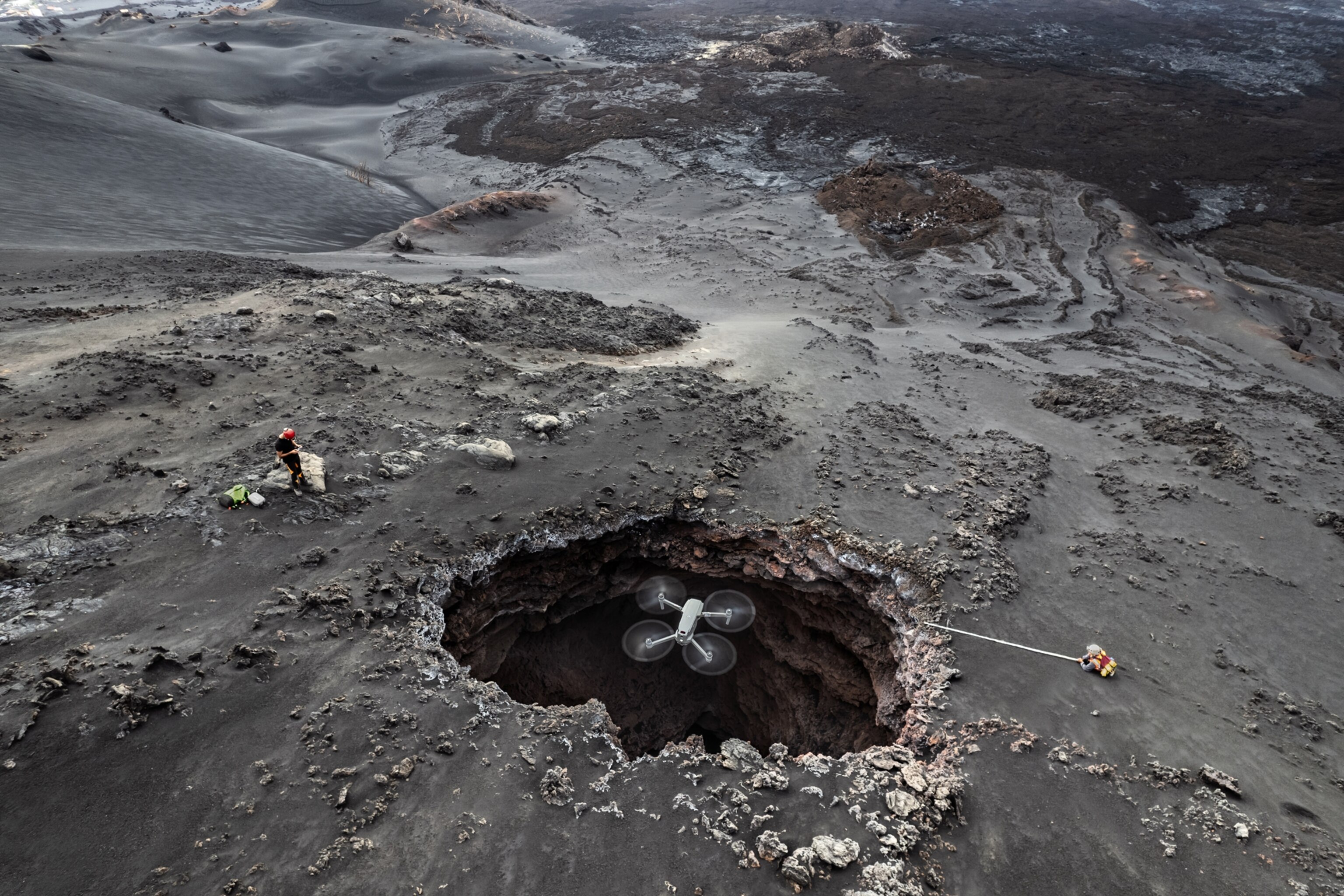 An aerial view showing a large hole amongst an ash-covered volcanic landscape. A drone measuring temperatures is situated such that it is framed by the gaping hole.