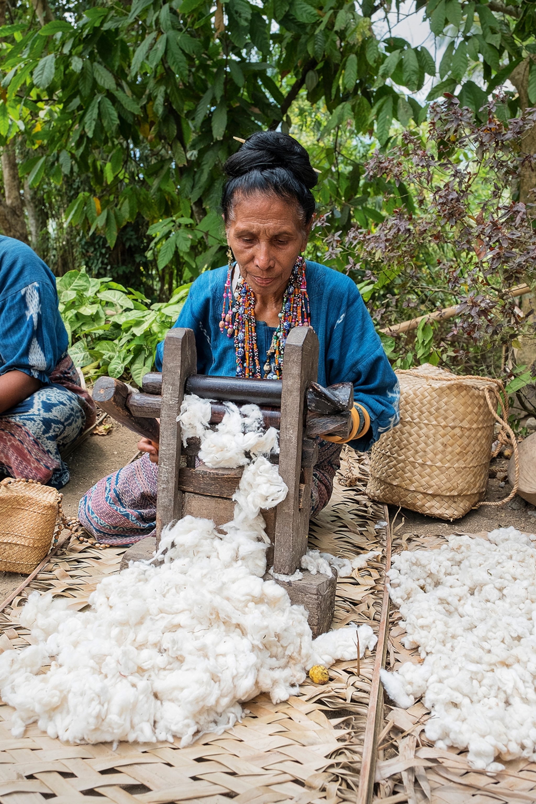 A local woman sitting a woven bamboo mat outside, spinning raw cotton.