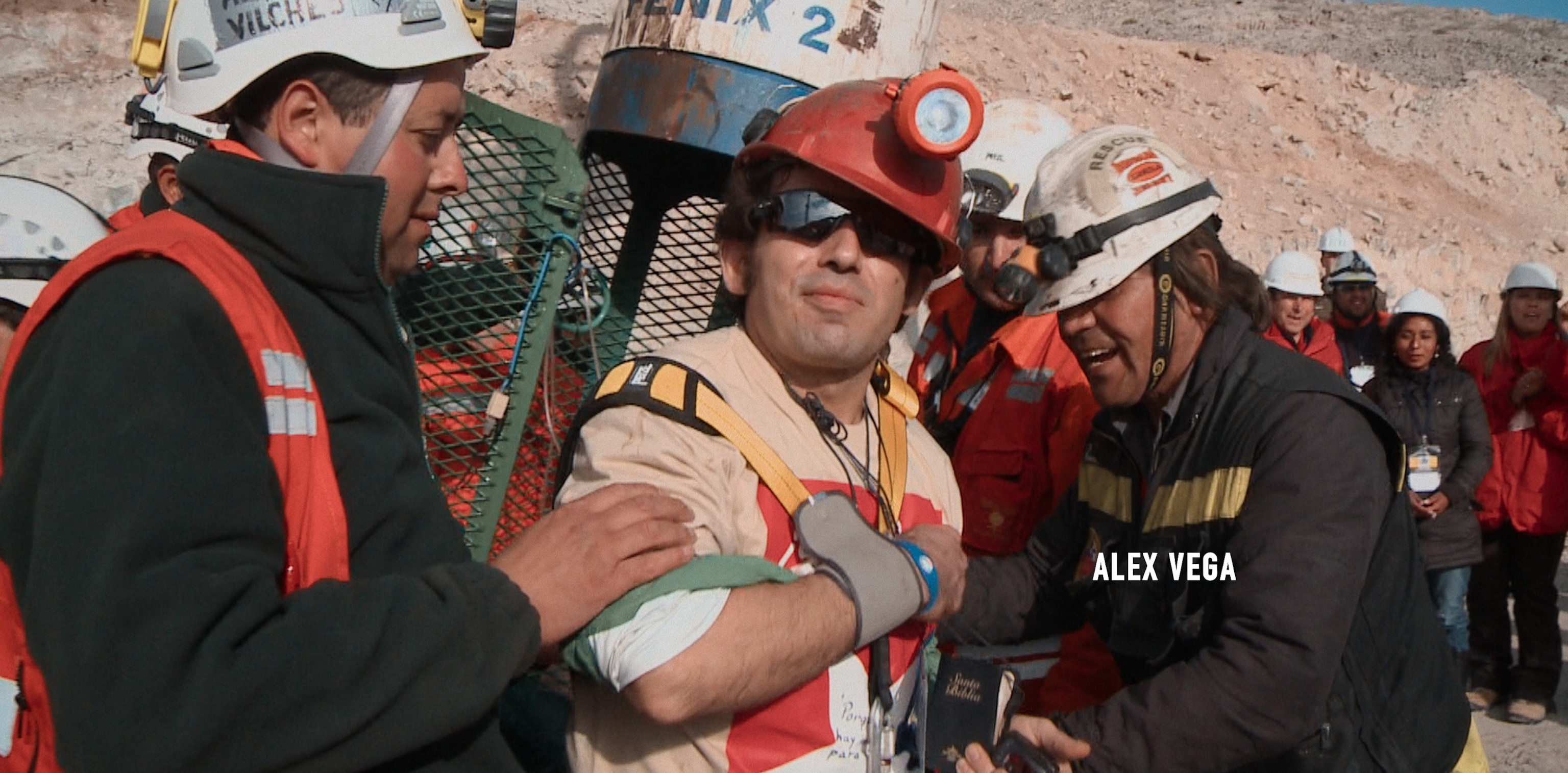 A man wearing an orange hardhat with a headlamp on it is helped up by two men in rescue gear on either side of him.